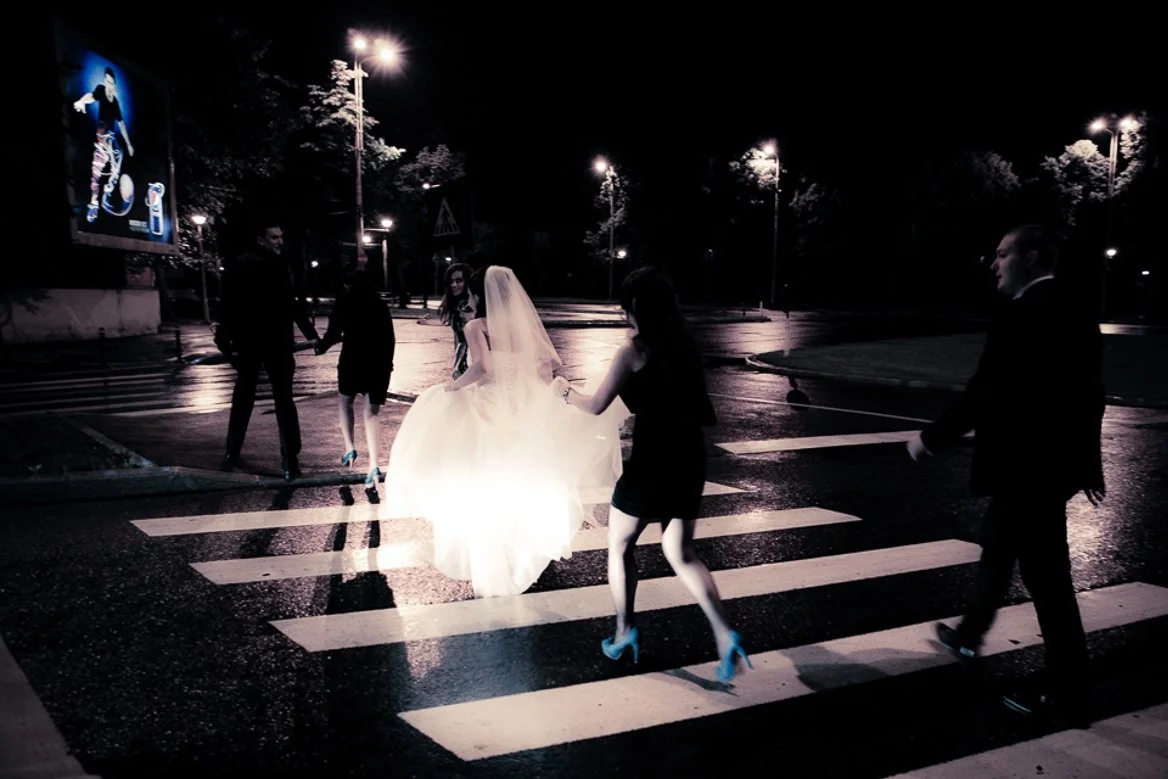 A bride in a white wedding dress and veil walking across a crosswalk at night, accompanied by four people dressed in black, holding her hands on a dark, rainy street with streetlights and trees.
