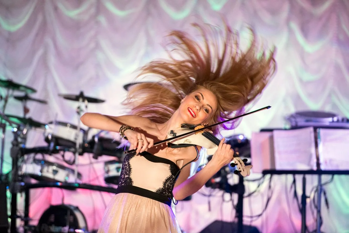 Violinist performing energetically on stage at a wedding with hair flipping, wearing a dress. Background shows musical equipment and a drum set.
