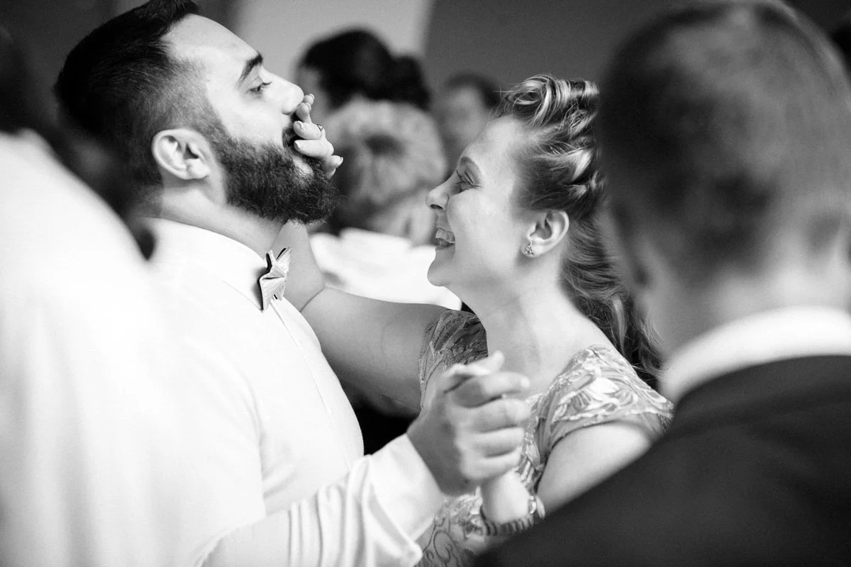 A black and white photograph of a smiling woman holding the face of a bearded man. They are dancing together in a crowded room. The man wears a dress shirt and bow tie.