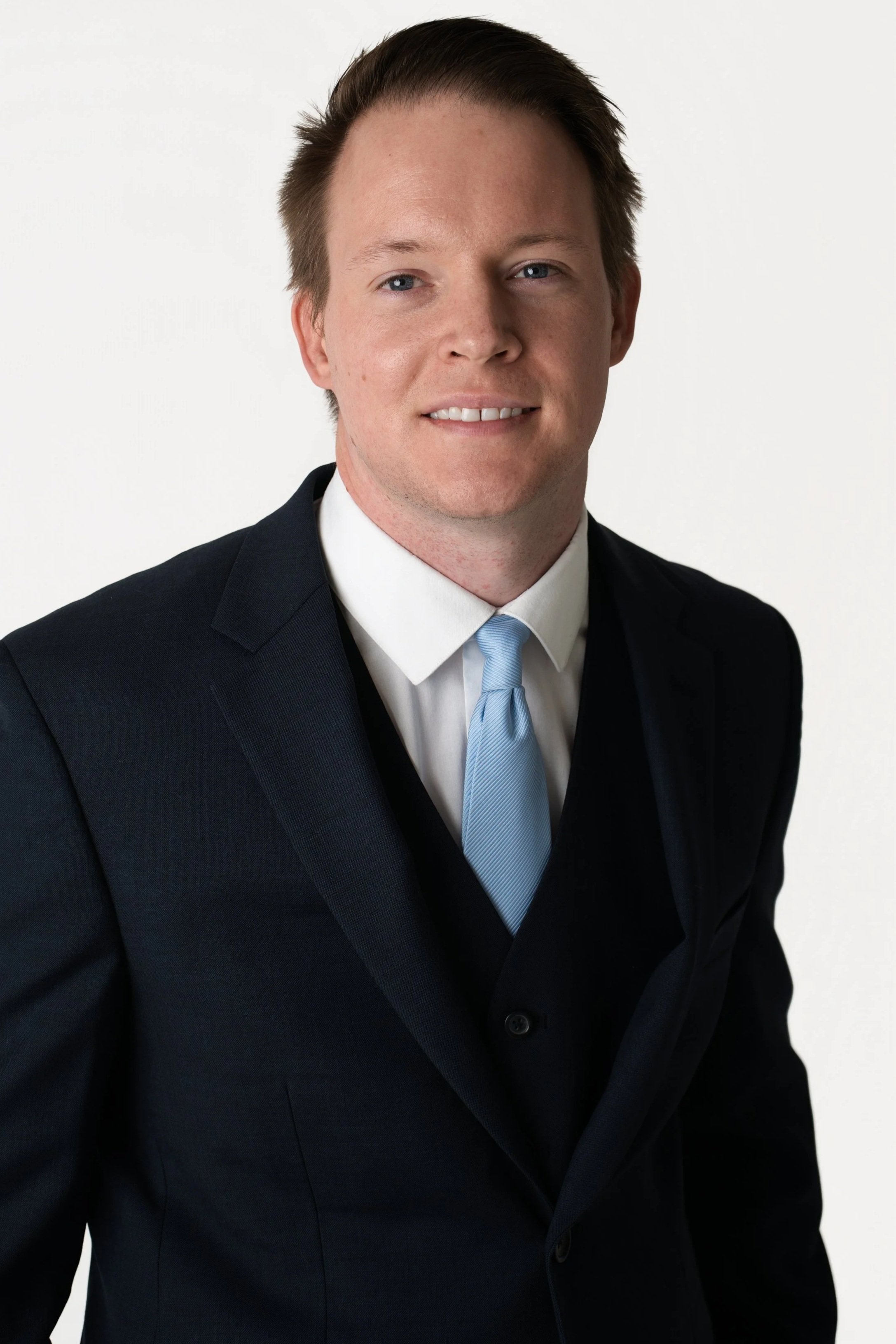 A young man with short brown hair, blue eyes, wearing a dark suit, white shirt, and red tie, smiling at the camera.