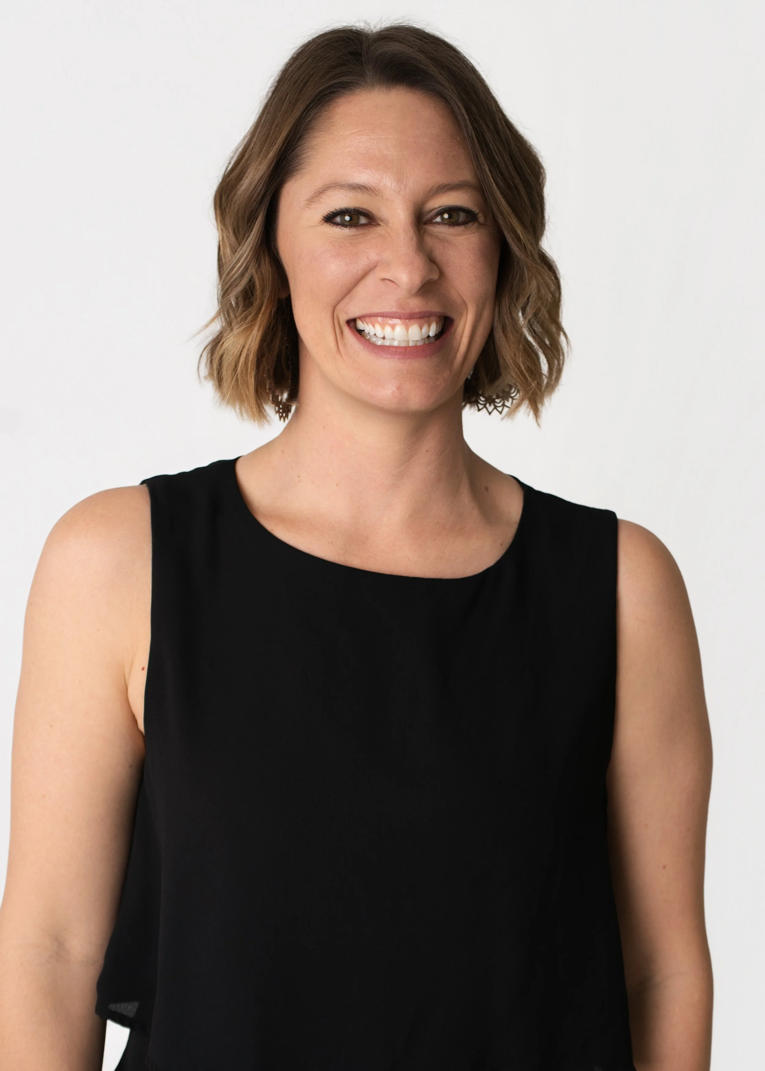 Portrait of a smiling woman with shoulder-length wavy brown hair, wearing a dark blazer and white top against a neutral background.