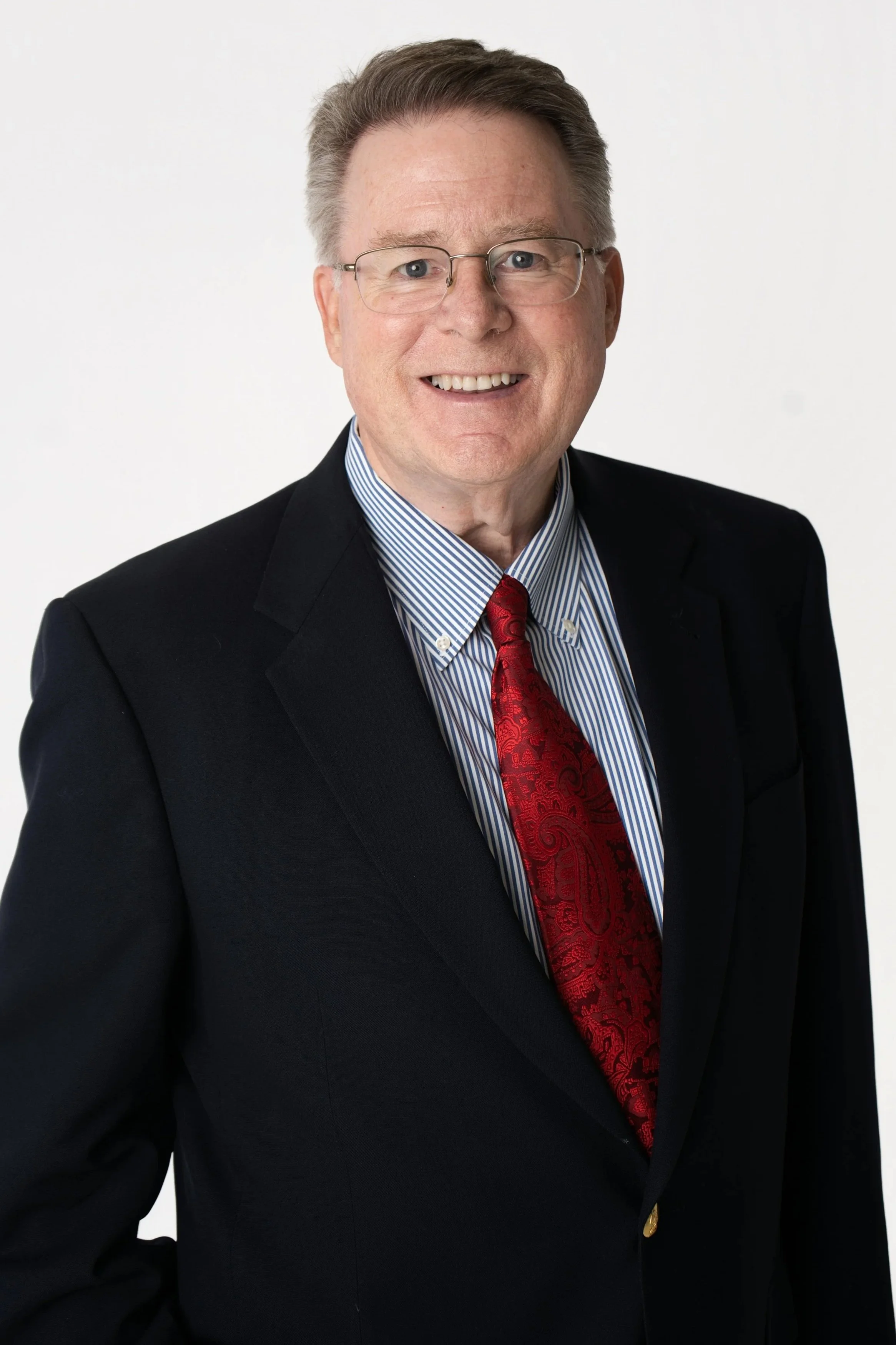 A middle-aged man with short brown hair, glasses, and light skin, dressed in a dark suit, white shirt, and striped tie, smiling against a plain background.