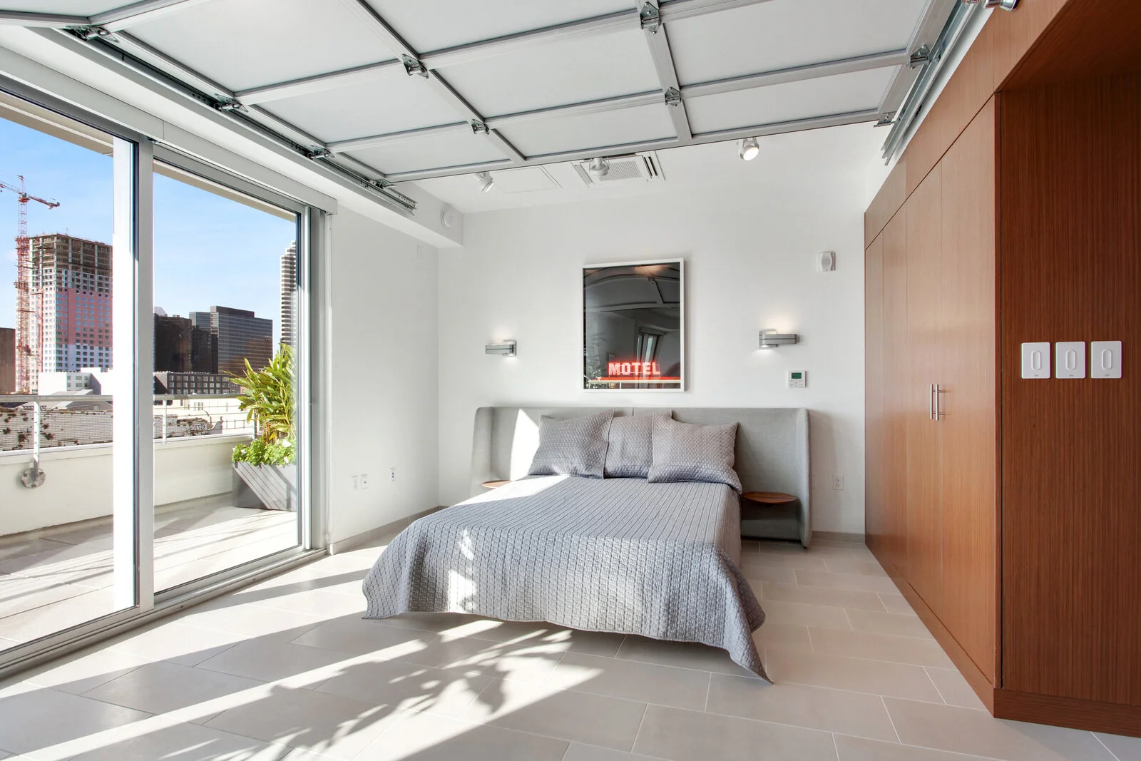  Luxury One Bedroom Apartments in New Orleans LA - The Garage - Furnished Bedroom with Tiled Floor, Sliding Garage Door on Ceiling, Grey Bed with Bedframe with Built-in Nightstands, Wooden Details and Large Closet, White Walls, and Floor-to-Ceiling W