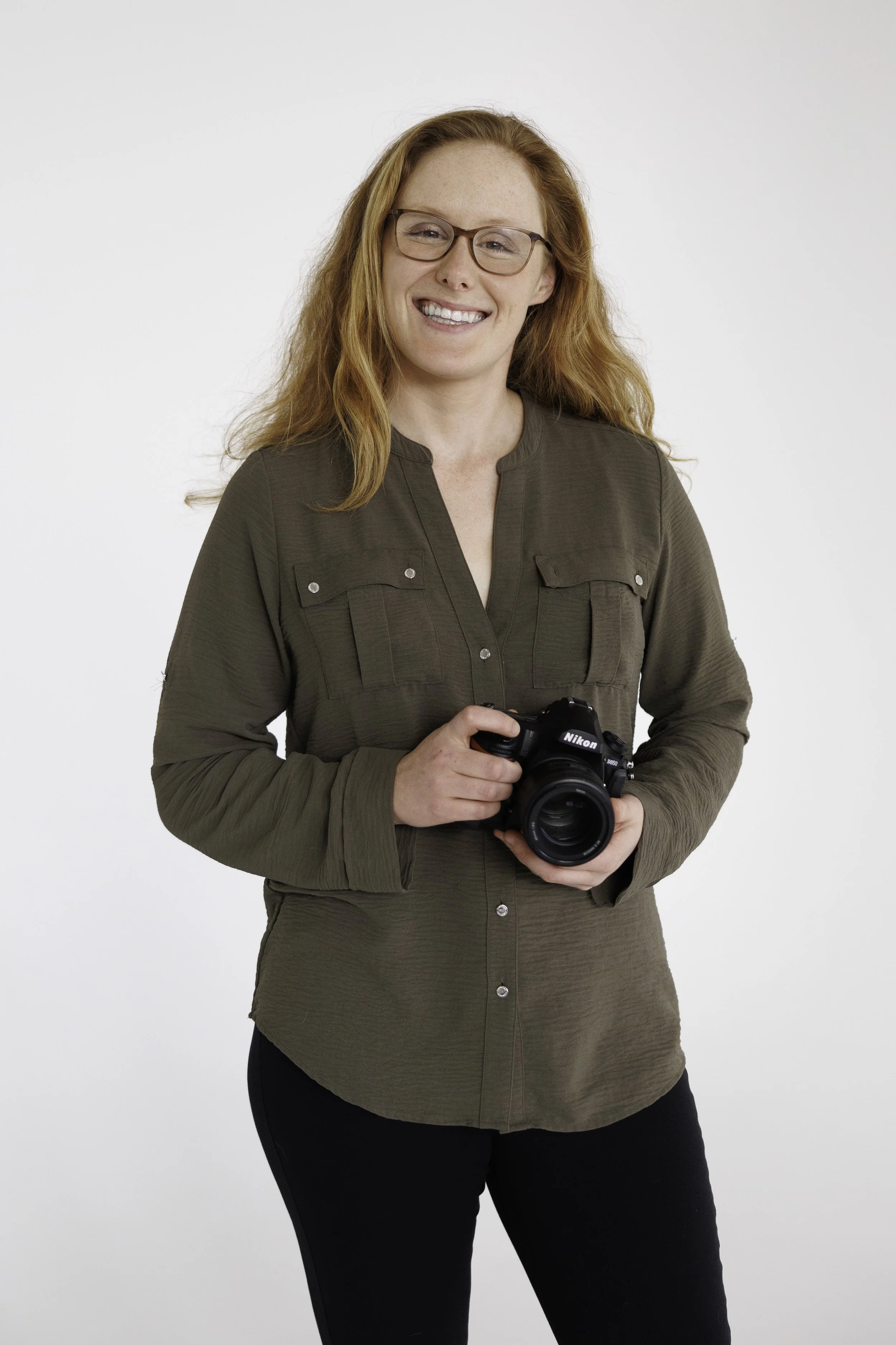 A woman with long red hair, glasses, and a smile holding a Nikon camera, standing in front of a plain white background wearing a dark green shirt and black pants.