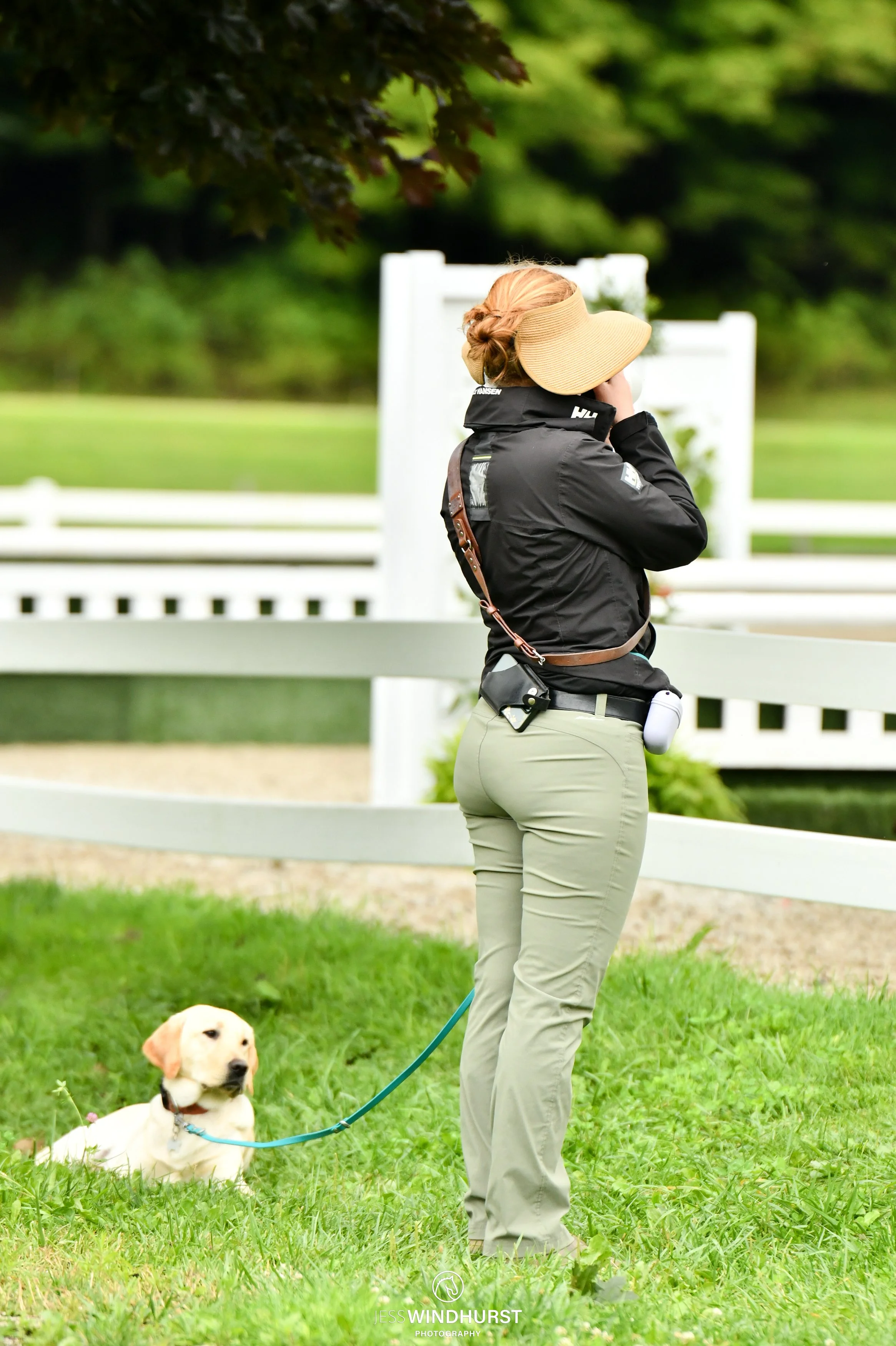A woman stands outdoors by a white fence, wearing a large straw hat, black jacket, and khaki pants, with a yellow Labrador retriever puppy sitting on green grass nearby.