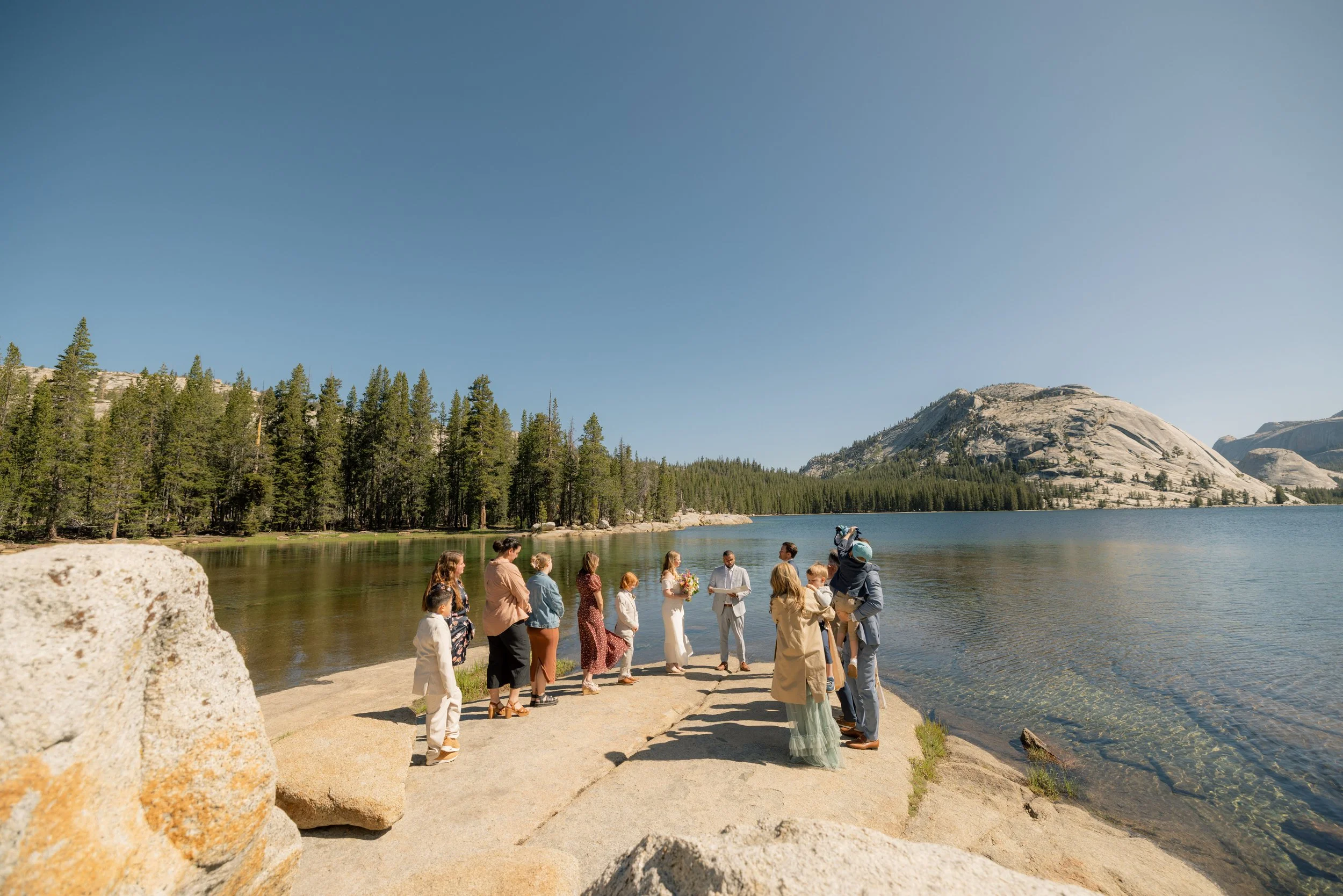 A wedding ceremony taking place outdoors on a rocky lakeshore surrounded by tall pine trees and mountains under a clear blue sky.