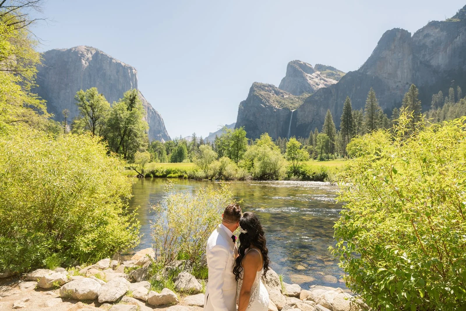 A couple in wedding attire standing closely together by a river in a lush green landscape with mountains and waterfalls in the background.