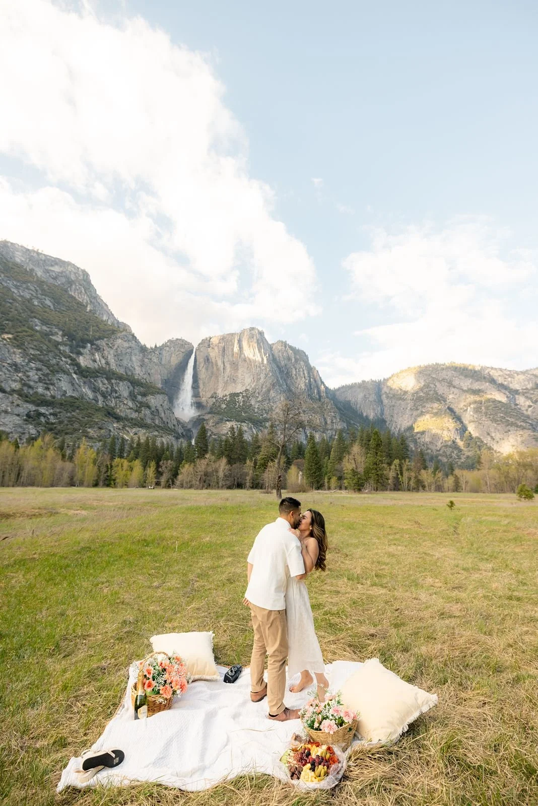Couple in white sharing a kiss in a green meadow with a massive Yosemite waterfall in the background.