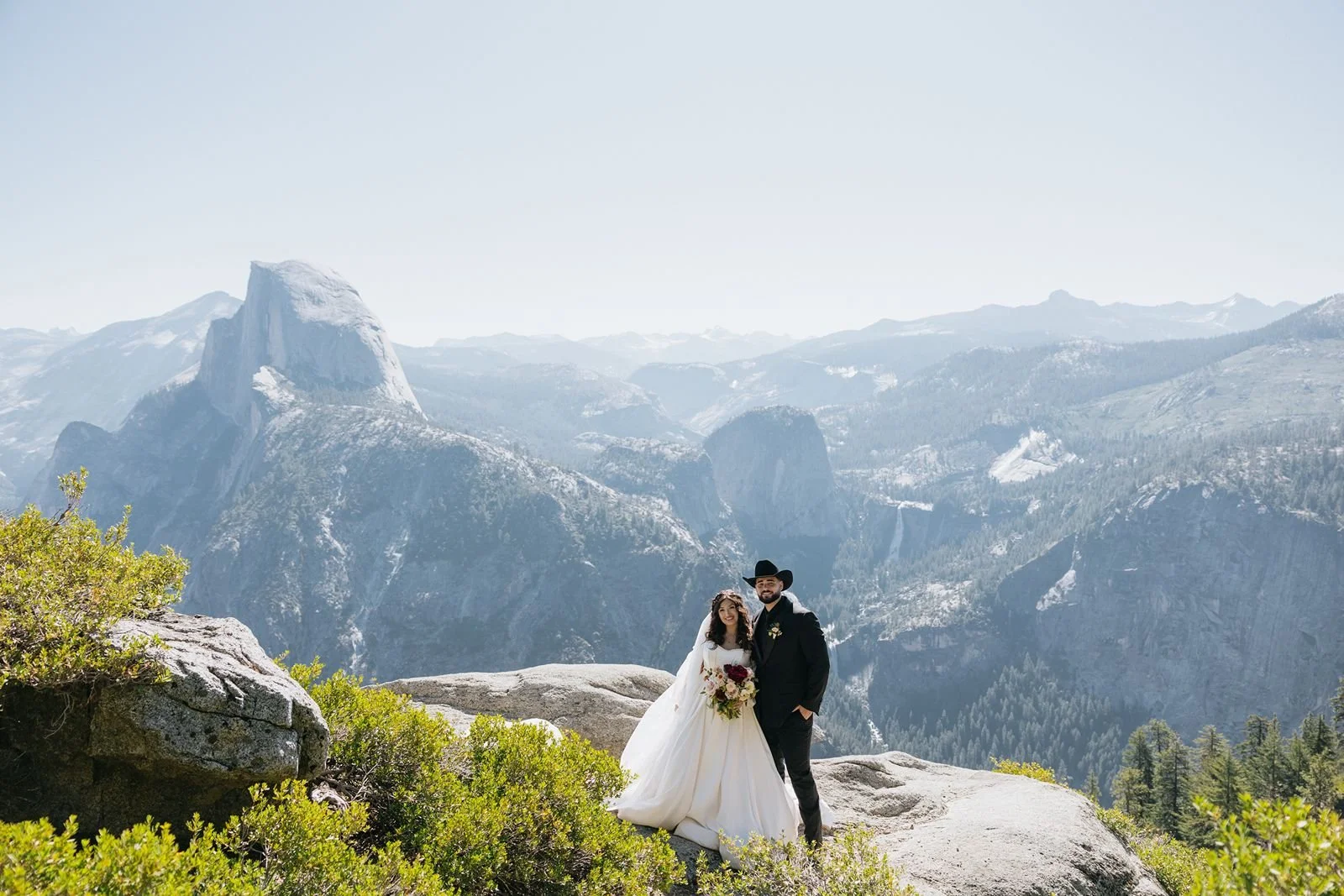 A bride and groom standing at Glacier Point in Yosemite during a dreamy sunrise, with soft morning light filtering through the mist over the valley and Half Dome.