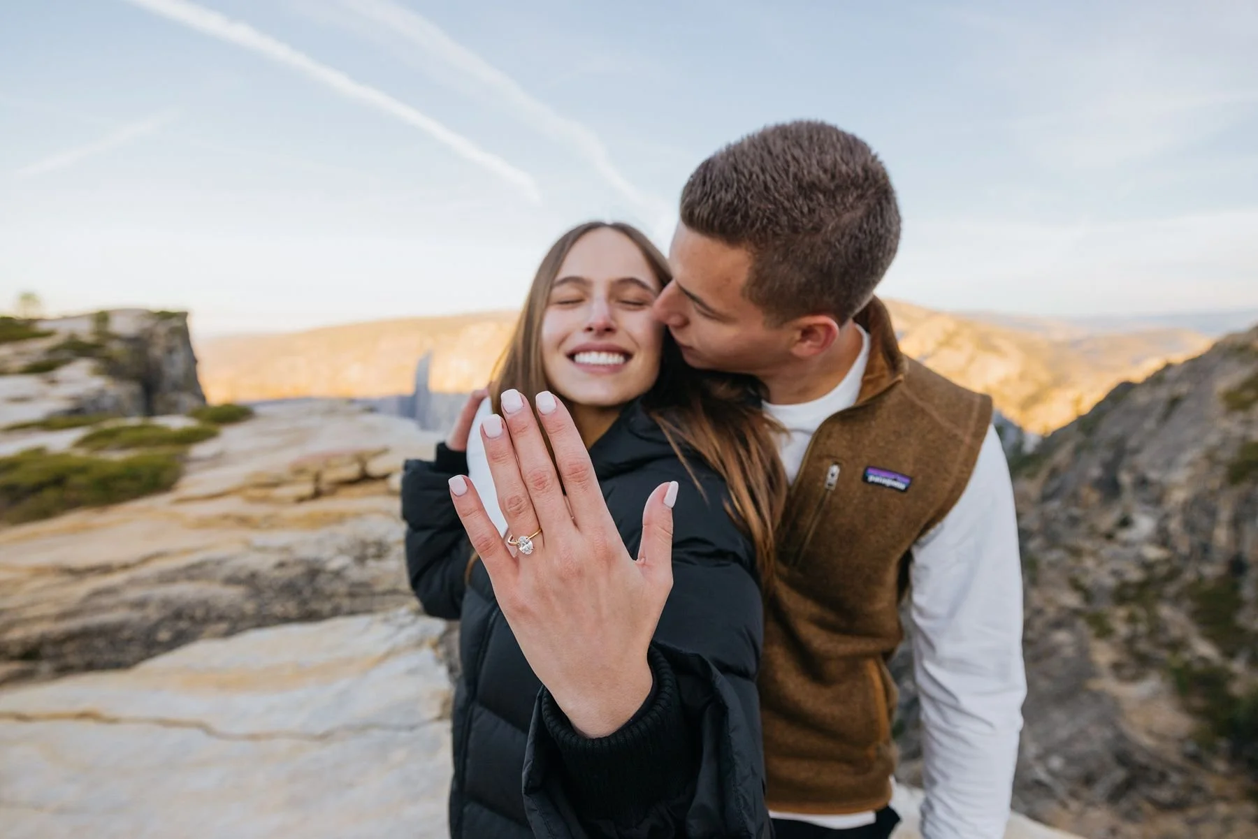 Happy woman showing her diamond engagement ring while her fiancé kisses her cheek at Taft Point, Yosemite.