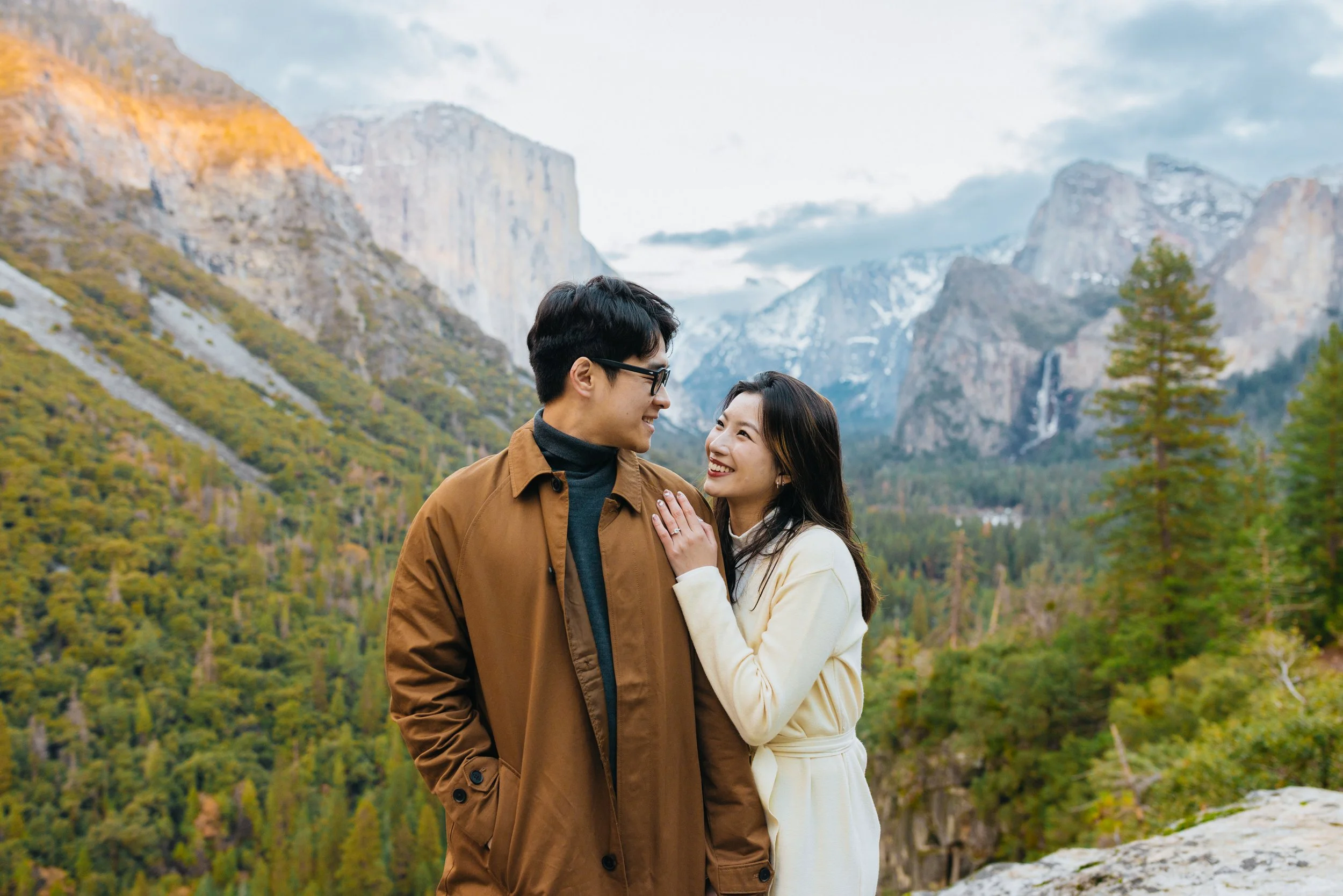 Newly engaged couple smiling at Tunnel View in Yosemite National Park with mountains and a cloudy sky.