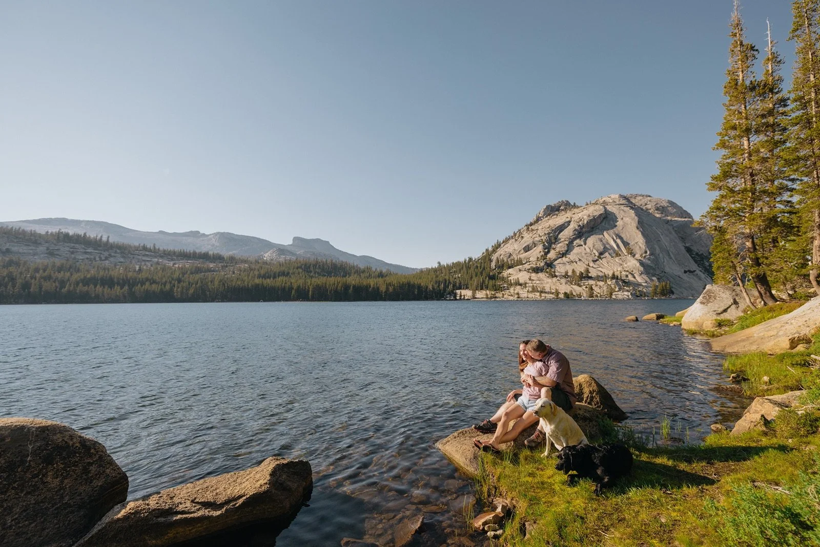 A couple with two dogs sitting on rocks at the edge of a lake with mountains and trees in the background.