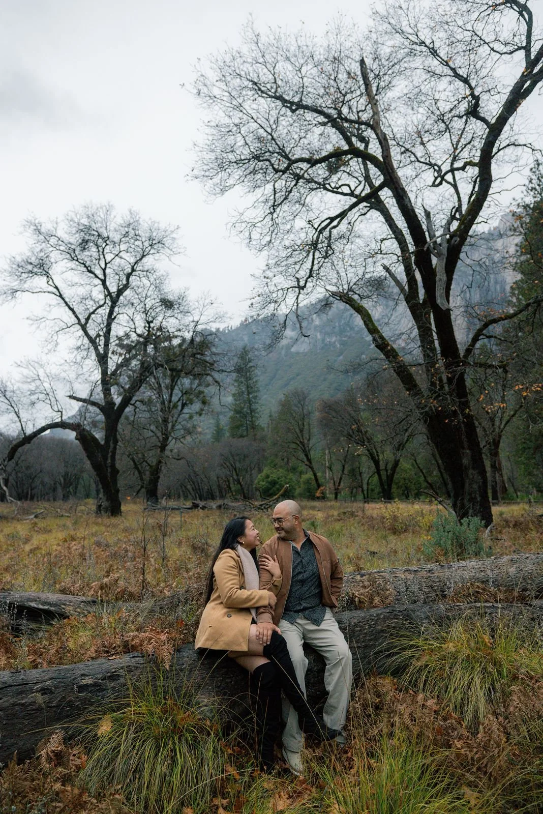 Engaged couple on a tree stump with cloudy Yosemite mountains in the background.
