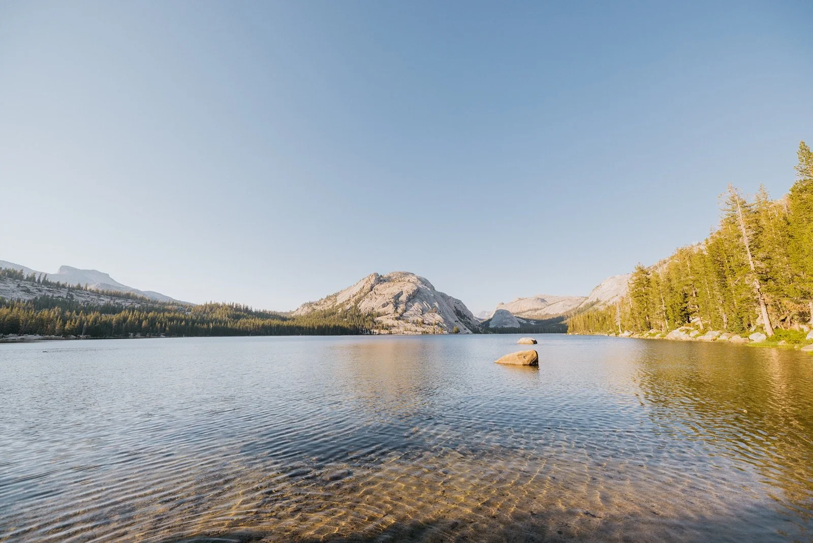 A serene mountain lake scene with calm water, surrounded by dense pine forests and rocky mountain peaks under a clear blue sky.