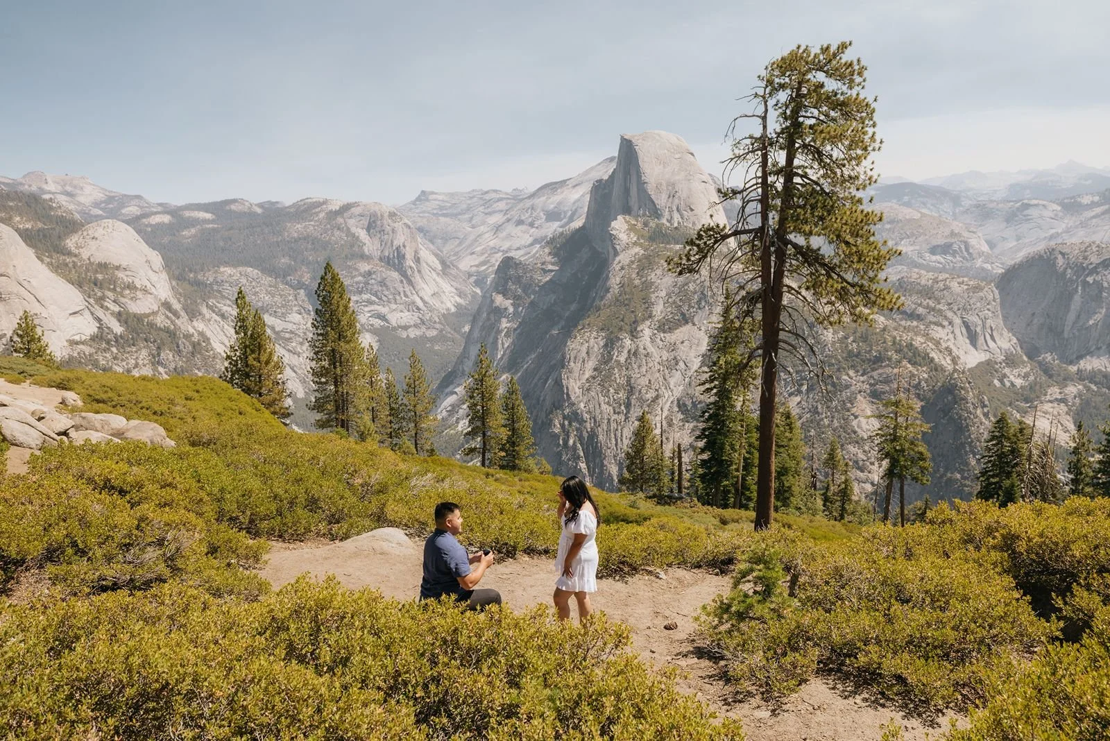 Man on one knee proposing to his fiancé at Glacier Point in Yosemite, surrounded by green trees and granite rocks.