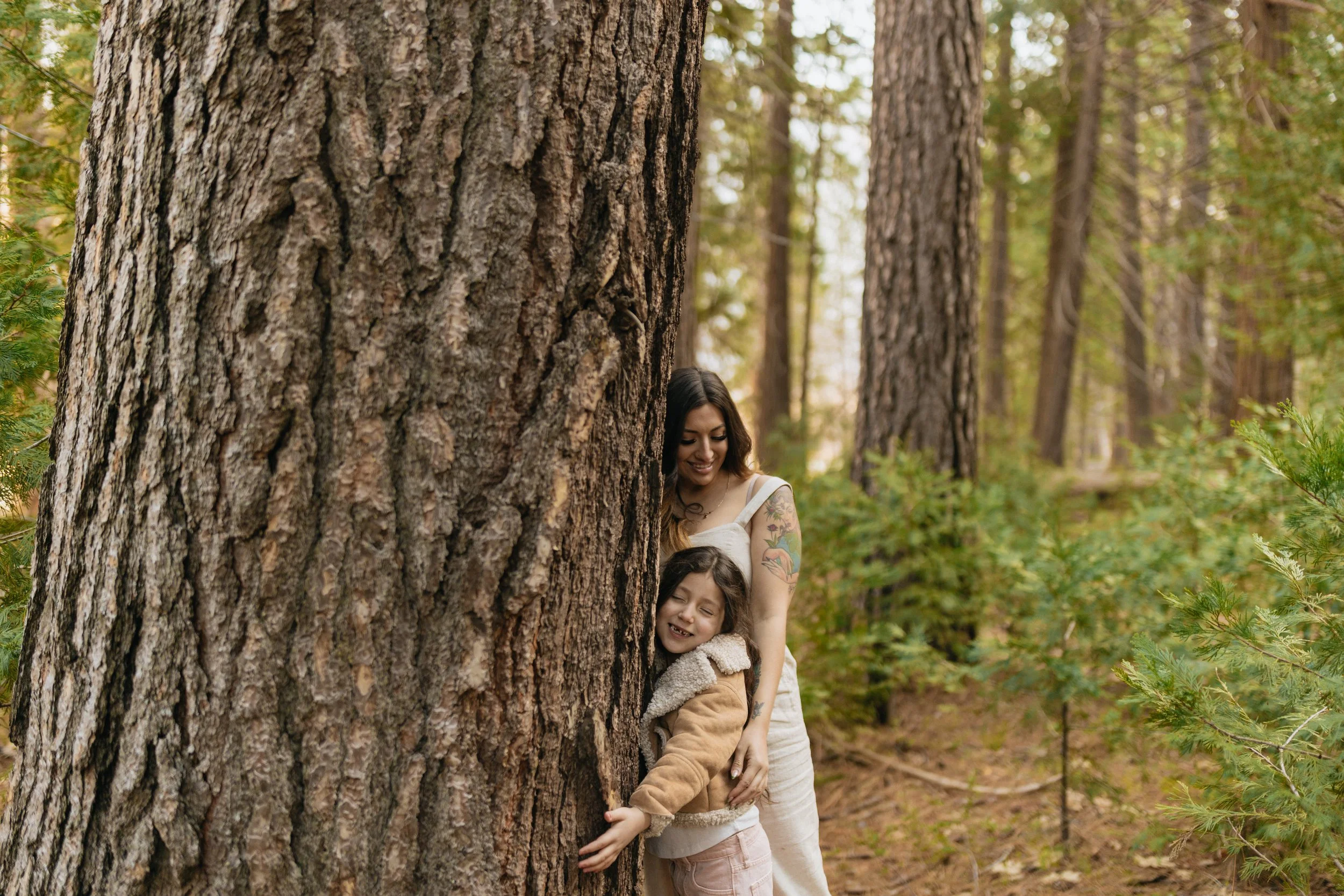 A woman and a girl smiling while hugging a large tree in the forest of Yosemite National Park.