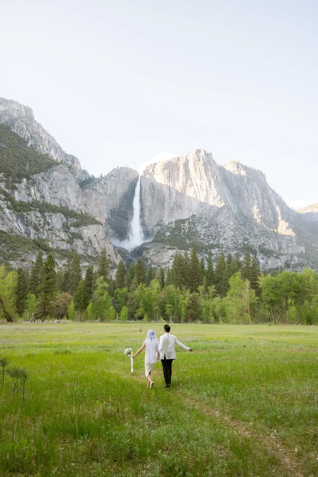 A bride and groom hold hands and walked towards a pouring waterfall in Yosemite