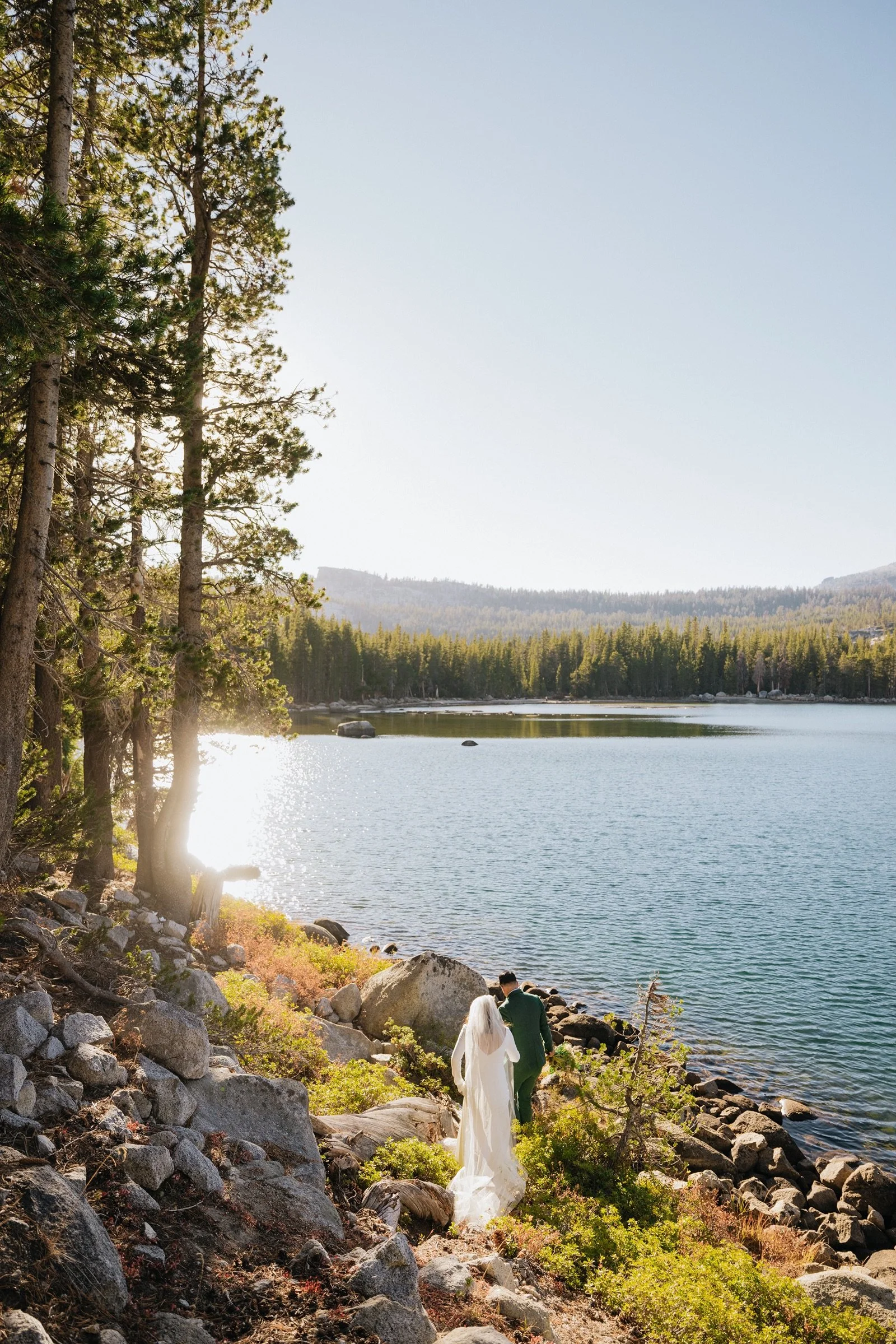A bride and groom walking along a rocky lakeshore surrounded by trees, with a mountain in the background and sunlight reflecting off the water.