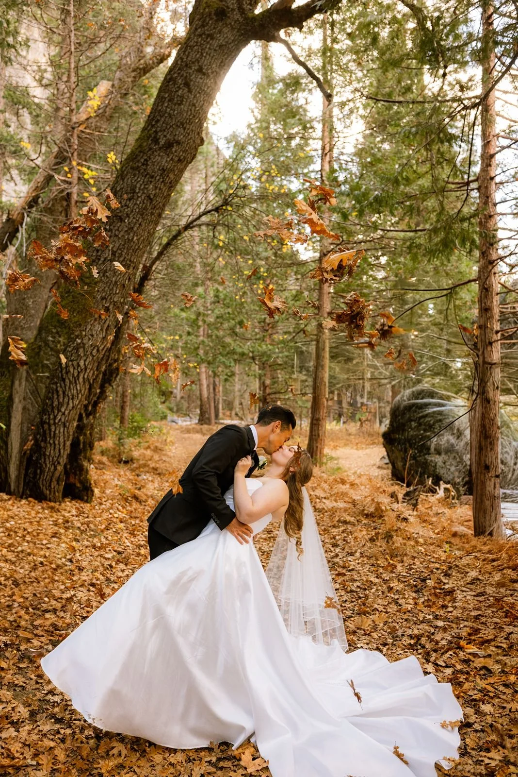 A bride and groom kiss with fall leaves falling from the trees
