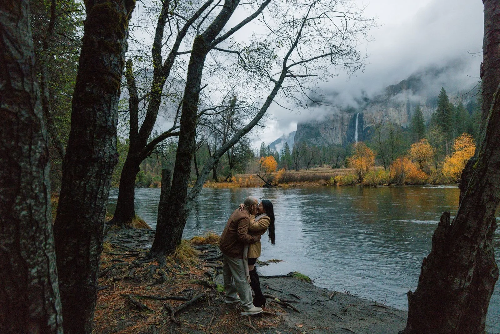 Couple embracing and kissing beside a flowing river with Yosemite mountain scenery in the background.