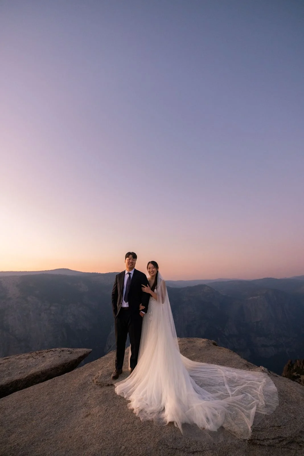 A bride and groom standing on a rocky ledge at sunset with a mountain landscape in the background. The bride is wearing a long white wedding dress with a train, and the groom is in a black suit with a white shirt and tie.