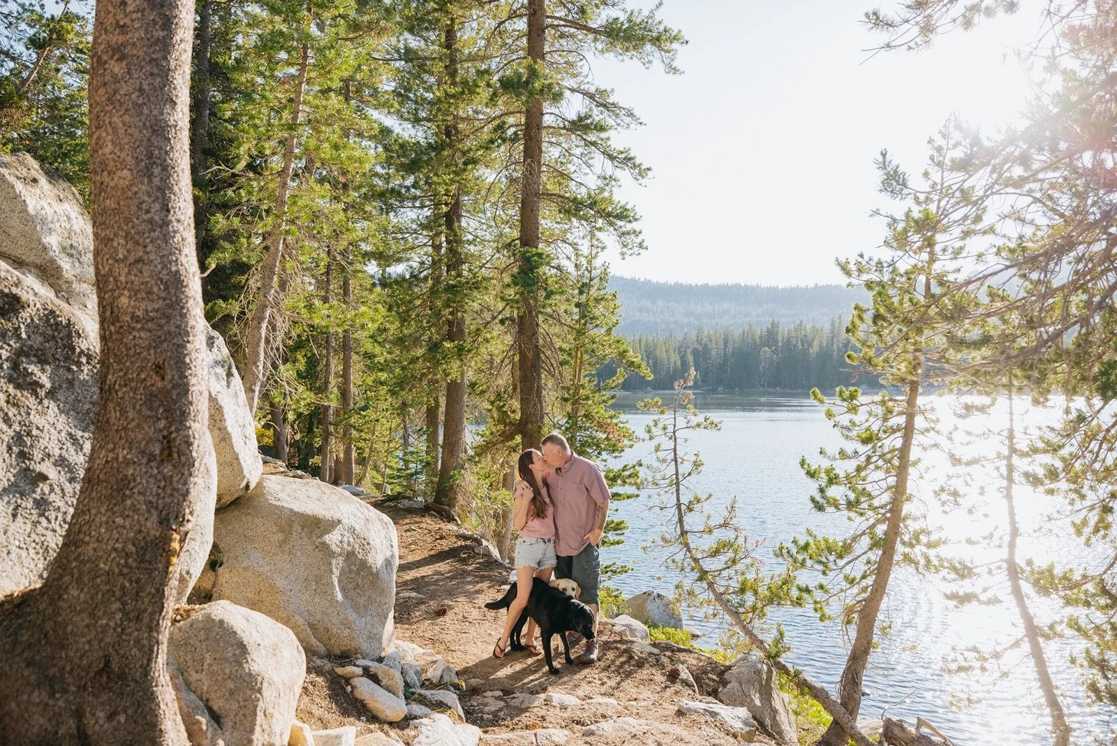 A couple sharing a kiss by a lake surrounded by trees with their two dogs, in a scenic nature setting during the daytime.