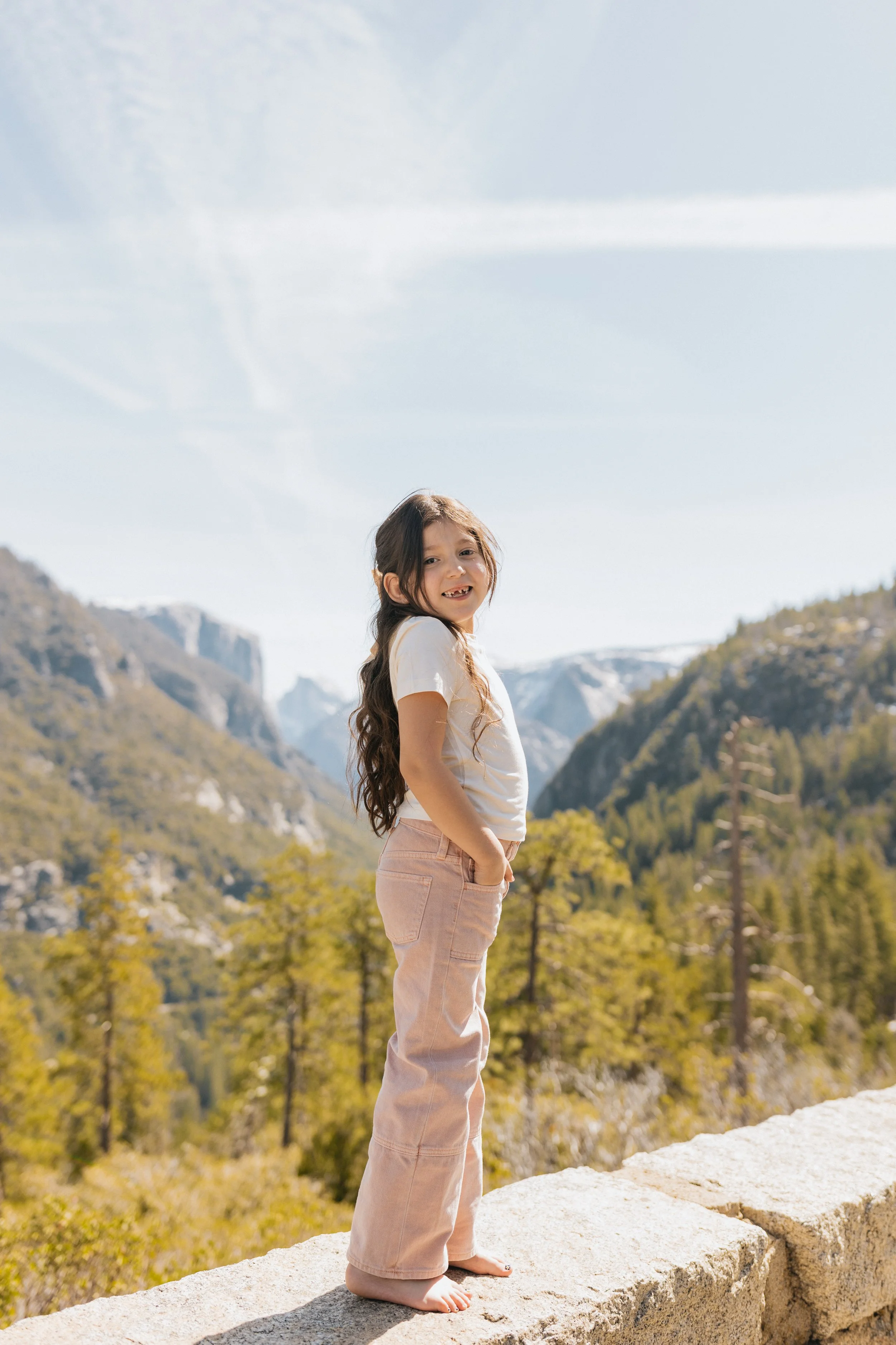 oung girl standing barefoot on a stone ledge at Tunnel View in Yosemite National Park with snow-capped peaks and green trees.
