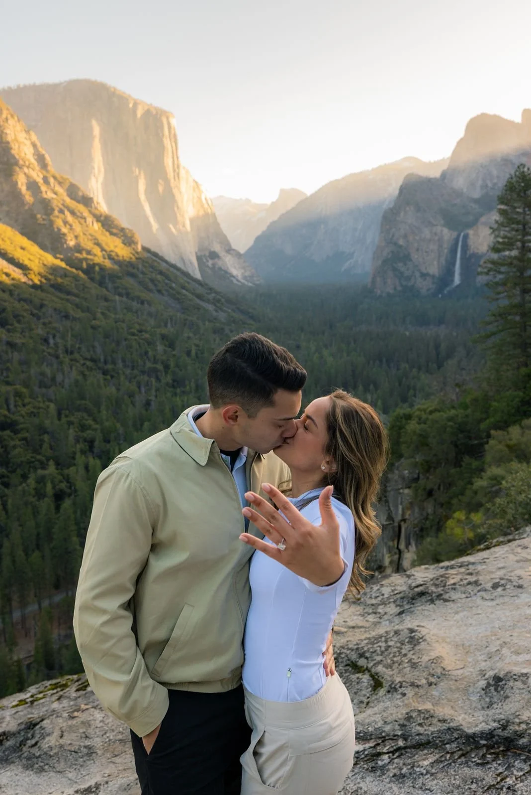 Newly engaged couple kissing and showing off a diamond ring with a Yosemite forest and waterfall background.