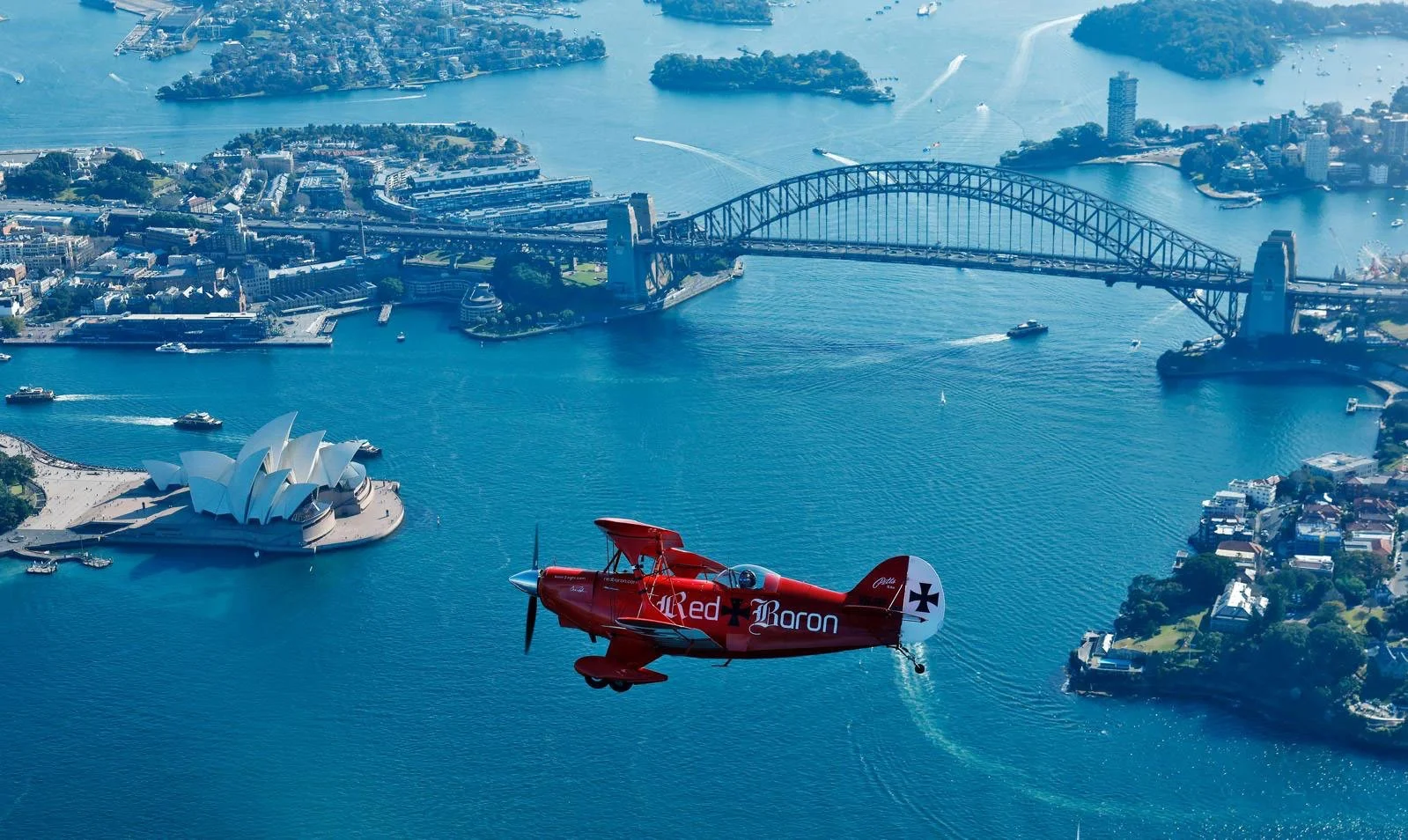 An aerial view of Sydney Harbour featuring the Sydney Opera House, Harbour Bridge, and a red vintage aircraft flying over the water.