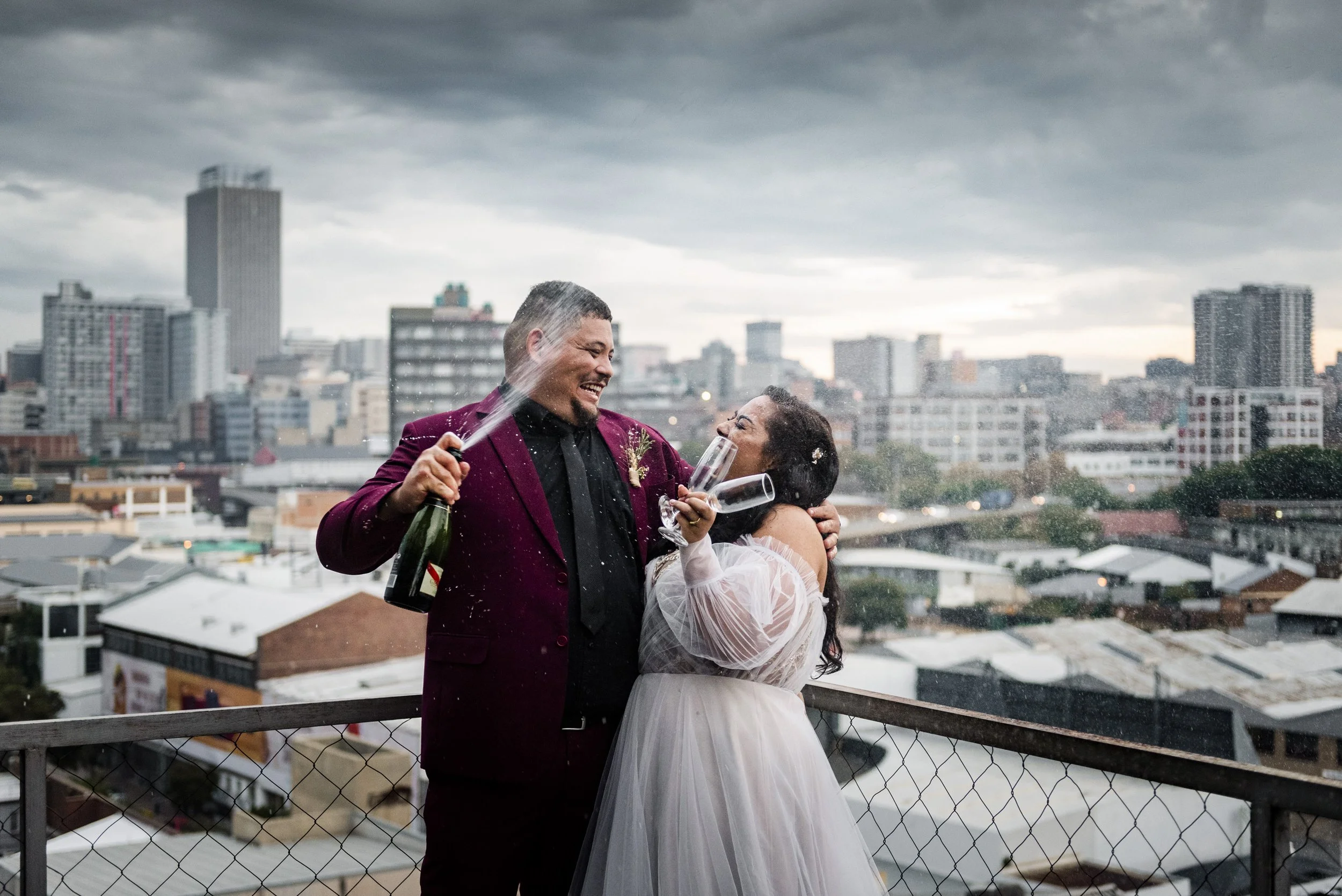 A couple celebrating on a rooftop with city buildings in the background, spraying champagne and smiling, amid gray cloudy skies. Photographed in the Johannesburg CBD. 