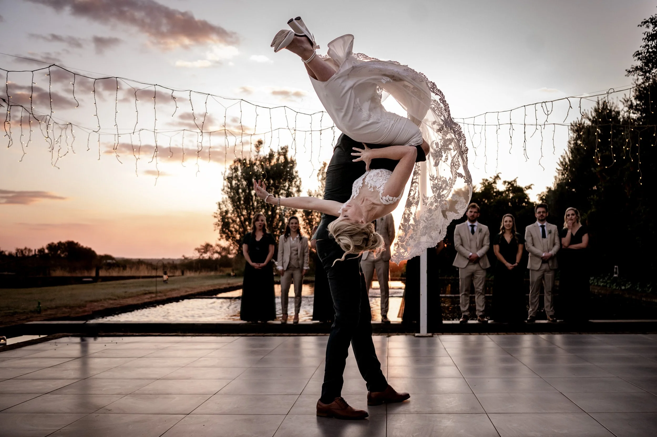 A bride and groom dancing during their outdoor wedding reception at sunset, with friends in the background and string lights overhead.