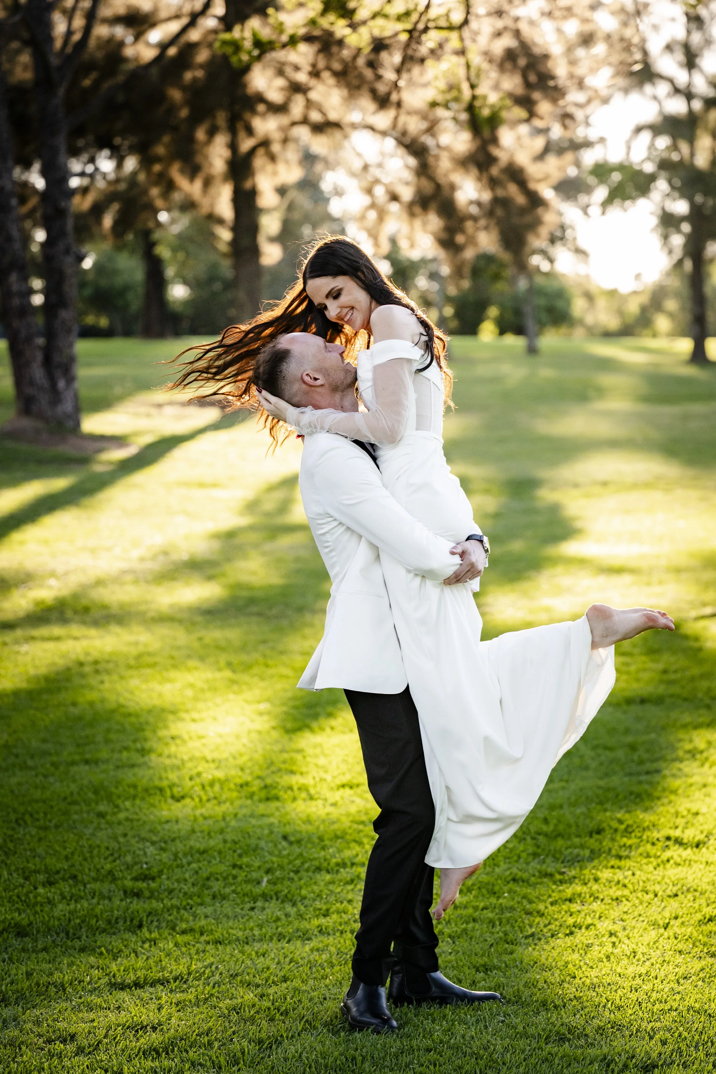 A couple in formal wedding attire enjoying time outdoors on a grassy field with trees in the background during sunset.