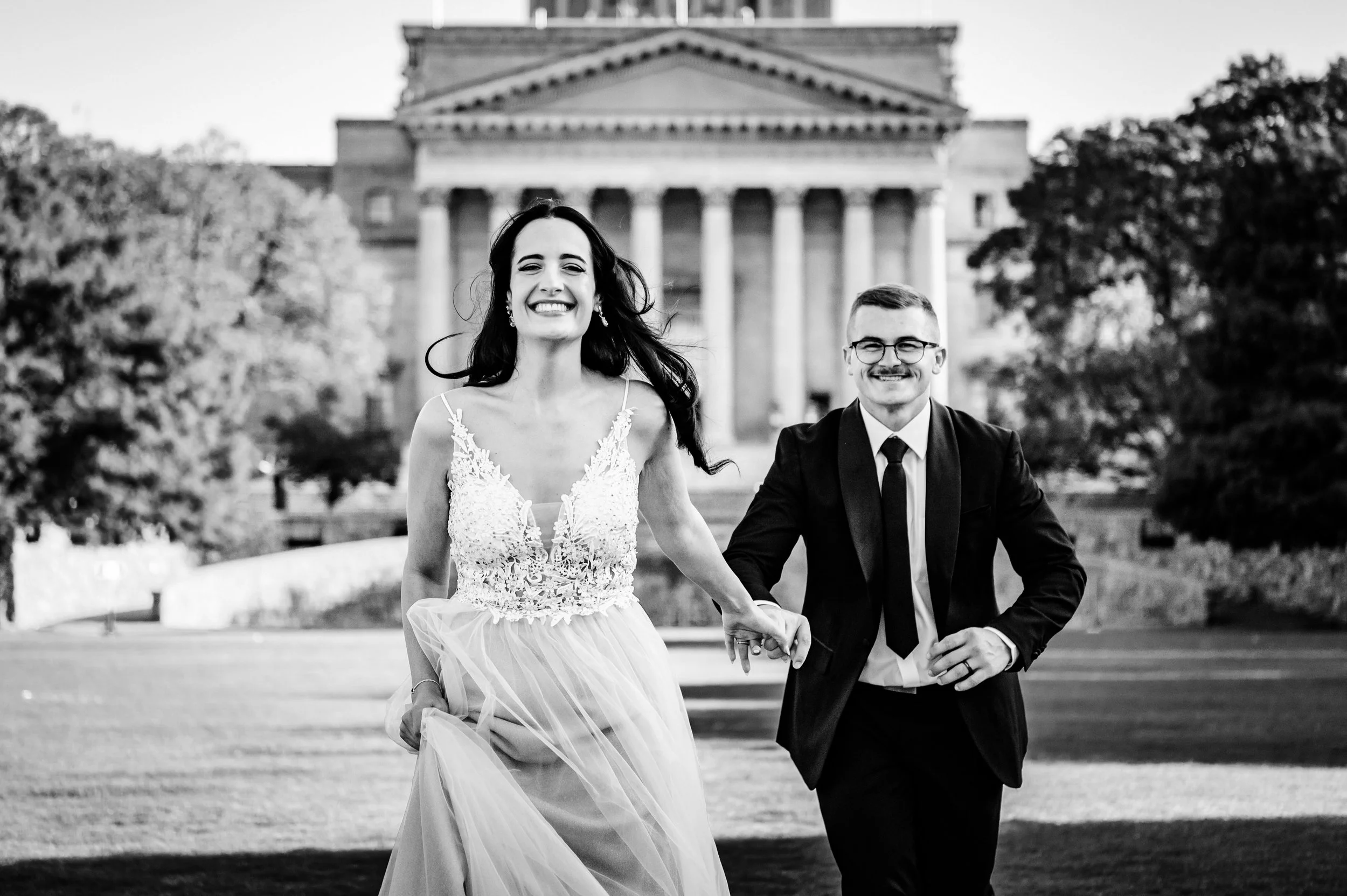 A black and white photo of a smiling bride and groom holding hands outdoors, with a large historic building with columns in the background.
