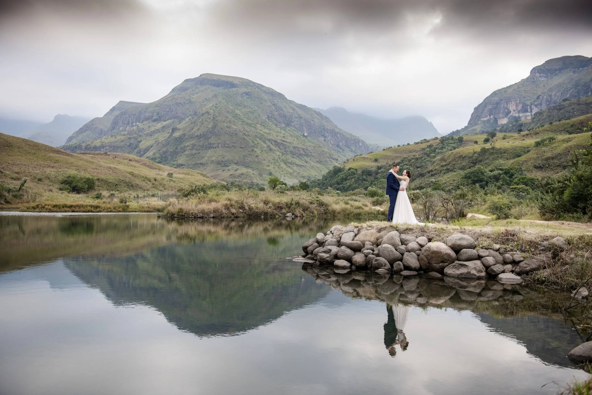 A couple in wedding attire standing by a calm body of water with mountains and overcast sky in the background. Photographed at Cathedral Peak in the Drakensberg Mountains. 