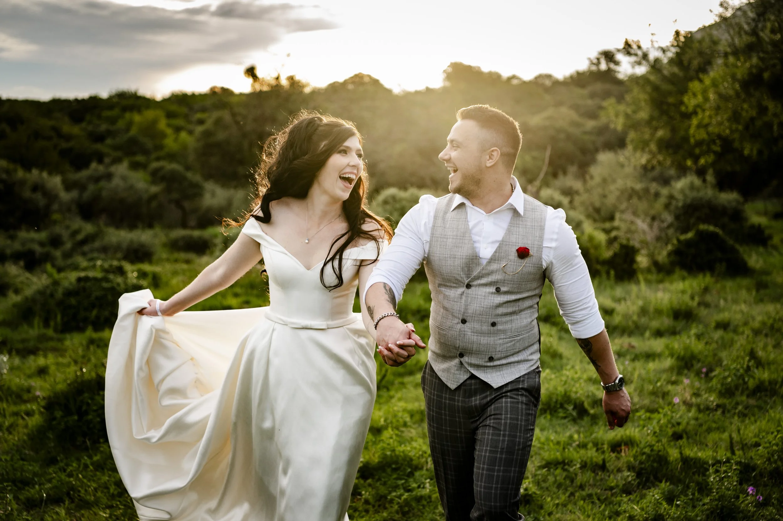 A couple holding hands and smiling while walking outdoors in a grassy area during sunset, with trees and cloudy sky in the background.