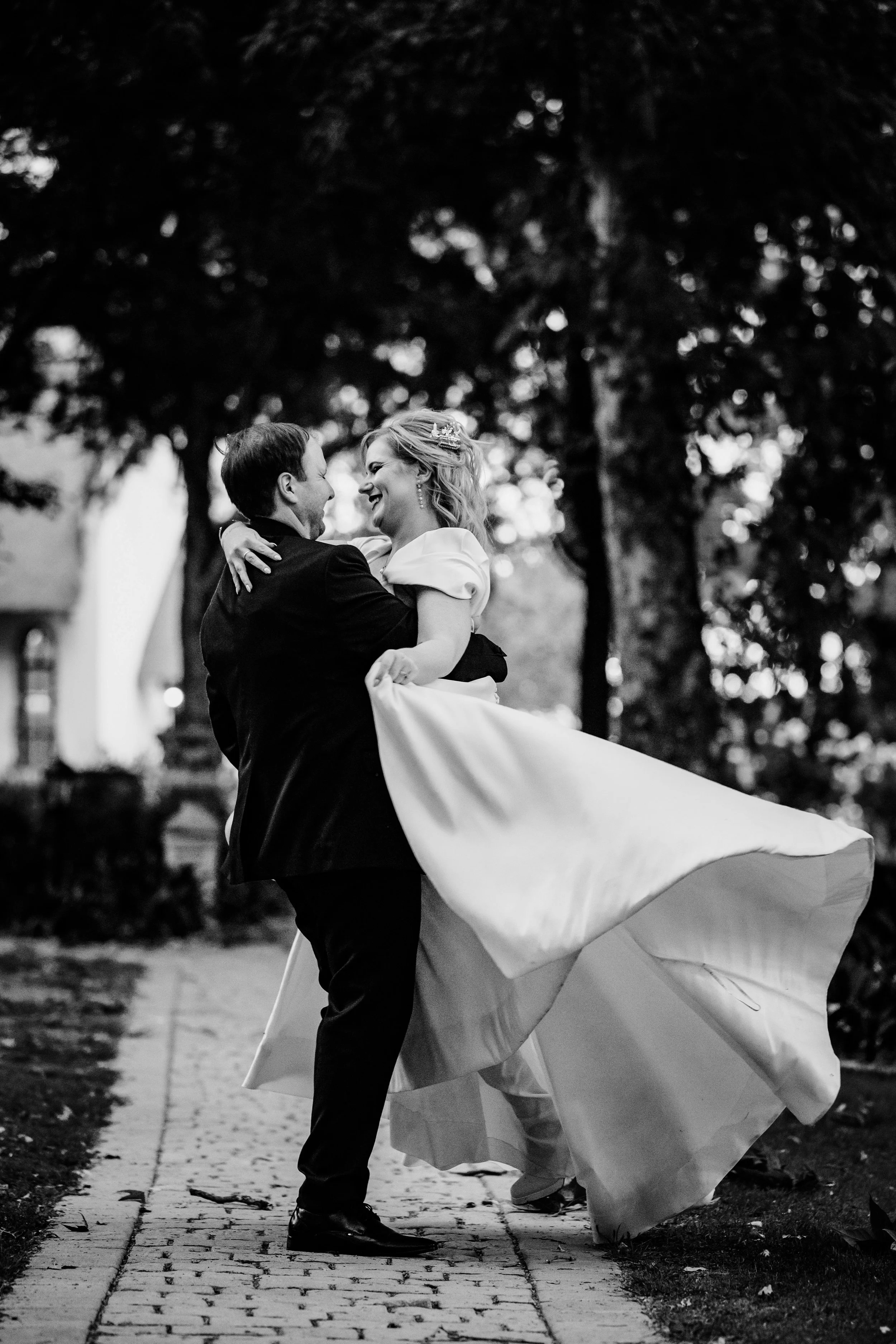 A couple dressed in formal wedding attire dancing outdoors on a cobblestone path, with trees and a building in the background, captured in black and white.
