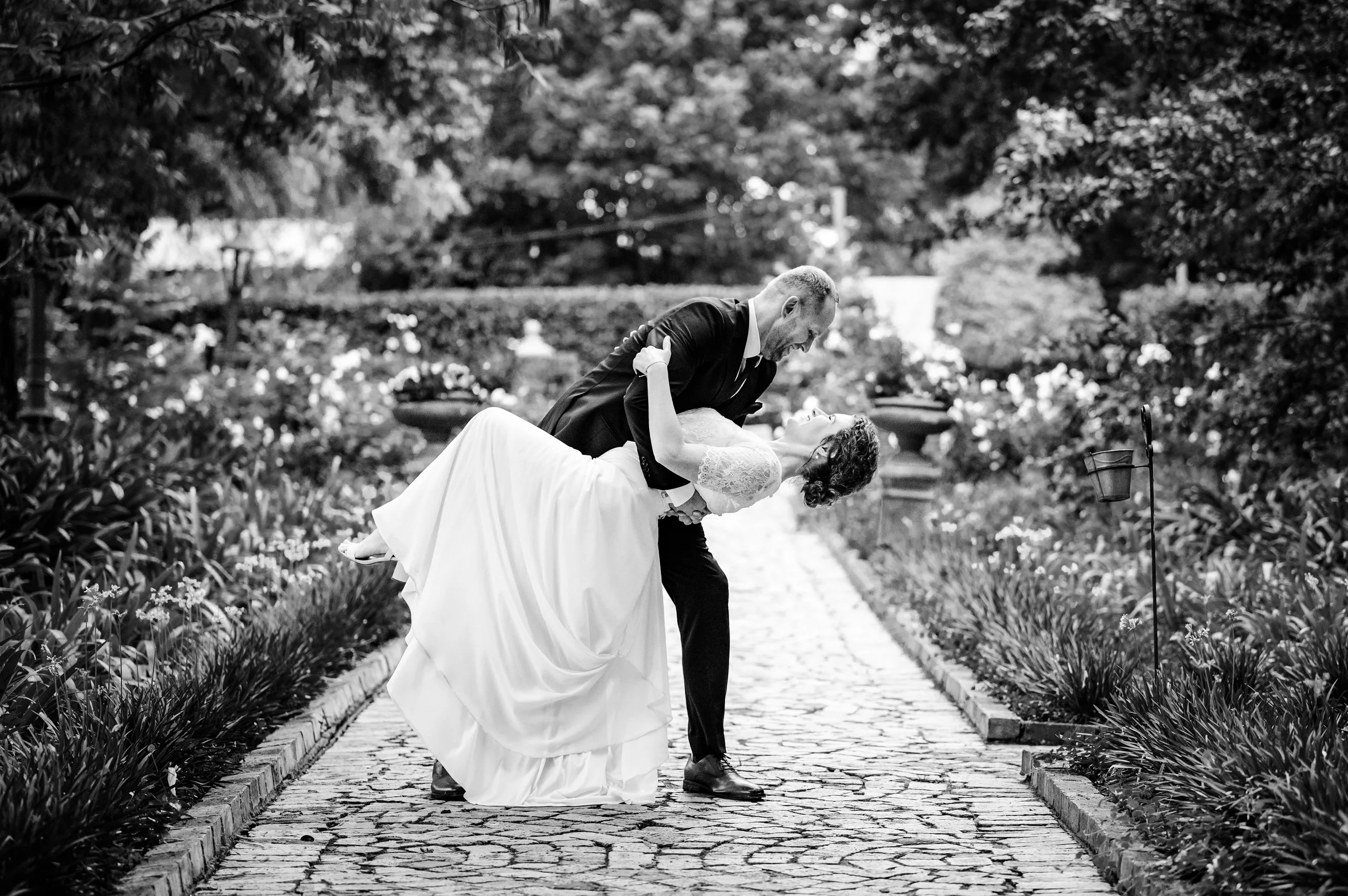 A black-and-white photograph of a bride and groom in a garden path, with the groom dipping the bride backwards. The bride is smiling and wearing a wedding gown, while the groom is dressed in a suit. The scene is surrounded by lush trees and plants.