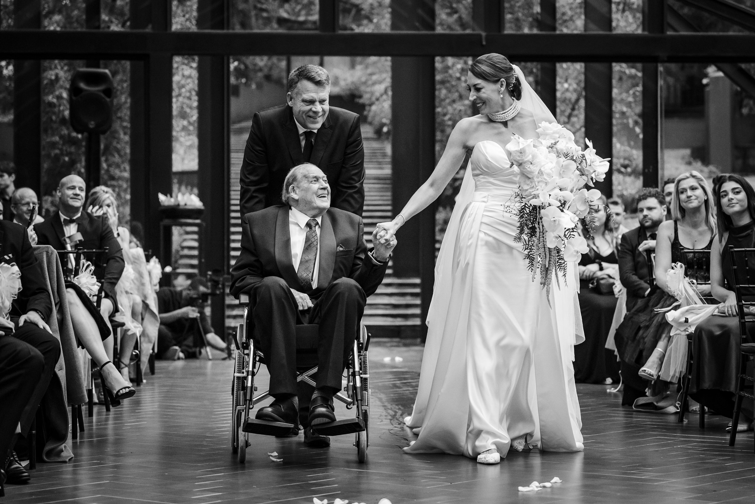 A bride in a wedding dress holding a bouquet and smiling, engaging with a man in a wheelchair during a wedding ceremony, with guests seated around. Photographed at Inimitable in Muldersdrift.