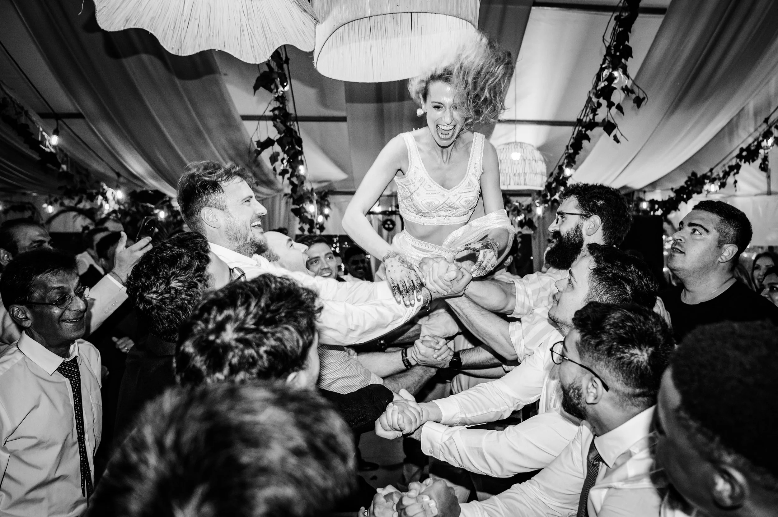A woman with curly hair celebrating on the shoulders of people at a party with decorations hanging from the ceiling. Photographed during a Jewish wedding called the Horas. 