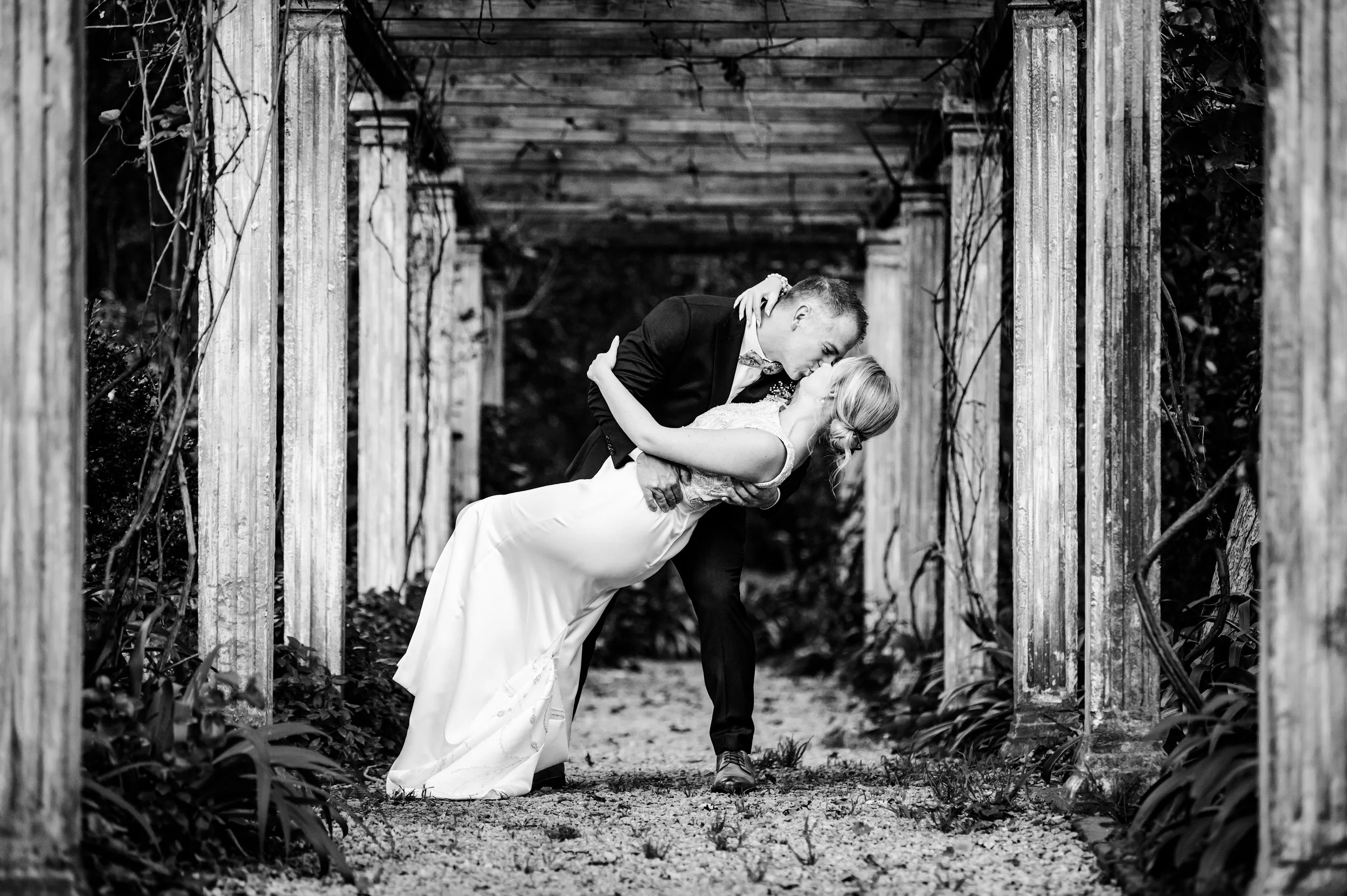 A black and white photo of a couple kissing, the man dips the woman in a dance pose on a pathway lined with wooden posts and vines.
