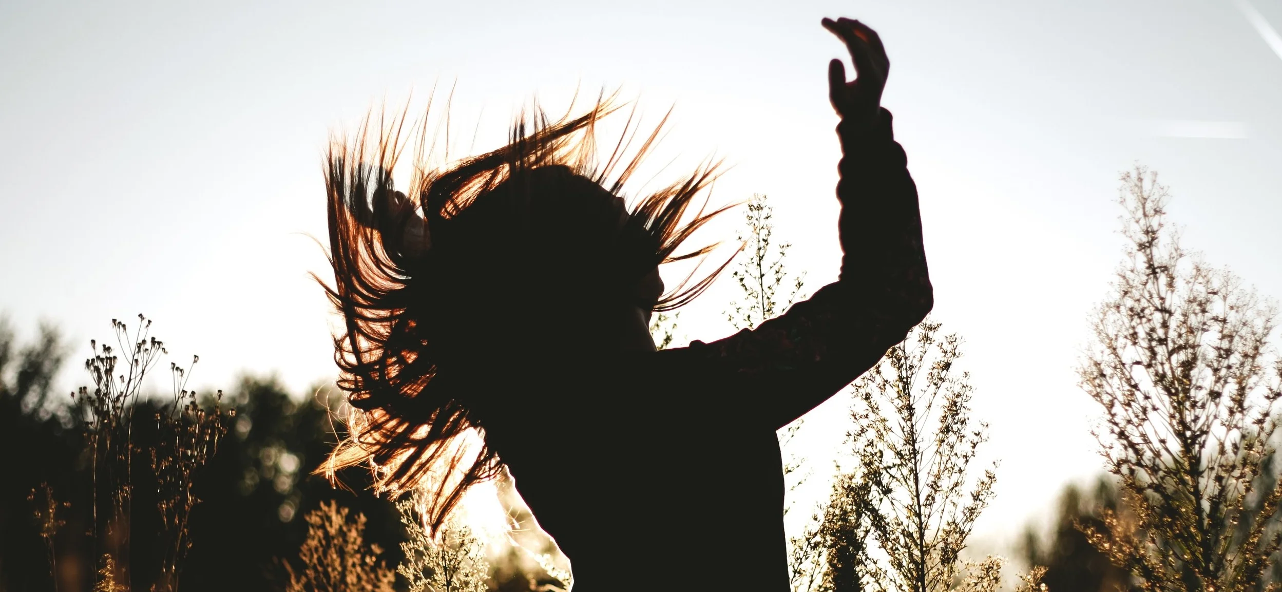 A girl in a meadow flipping her hair back. She is silhouetted by the sun behind her. You too can feel this good about your hair!