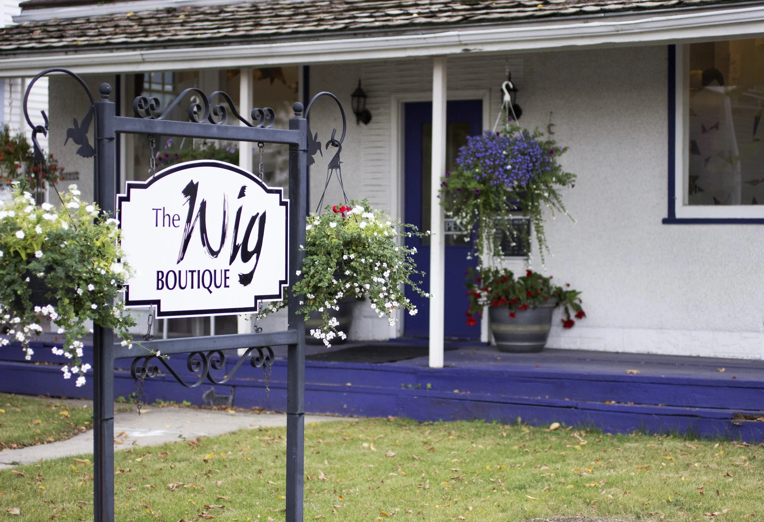 The outside of the shop. The Wig Boutique is inside a white house with a purple porch and door. There is a sign on the front yard.