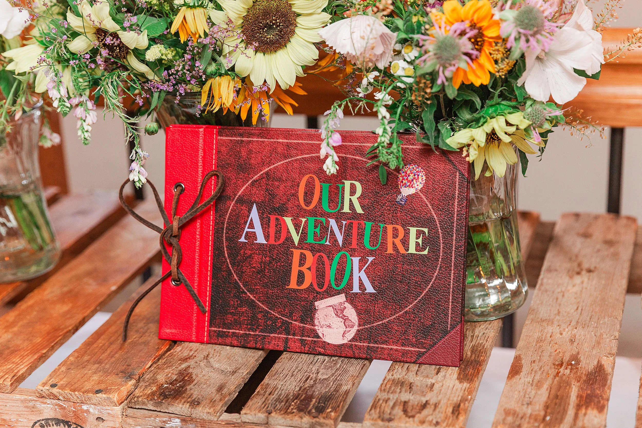 A rust coloured wedding guest book with our adventure book written on the front propped up against a display of wedding flowers