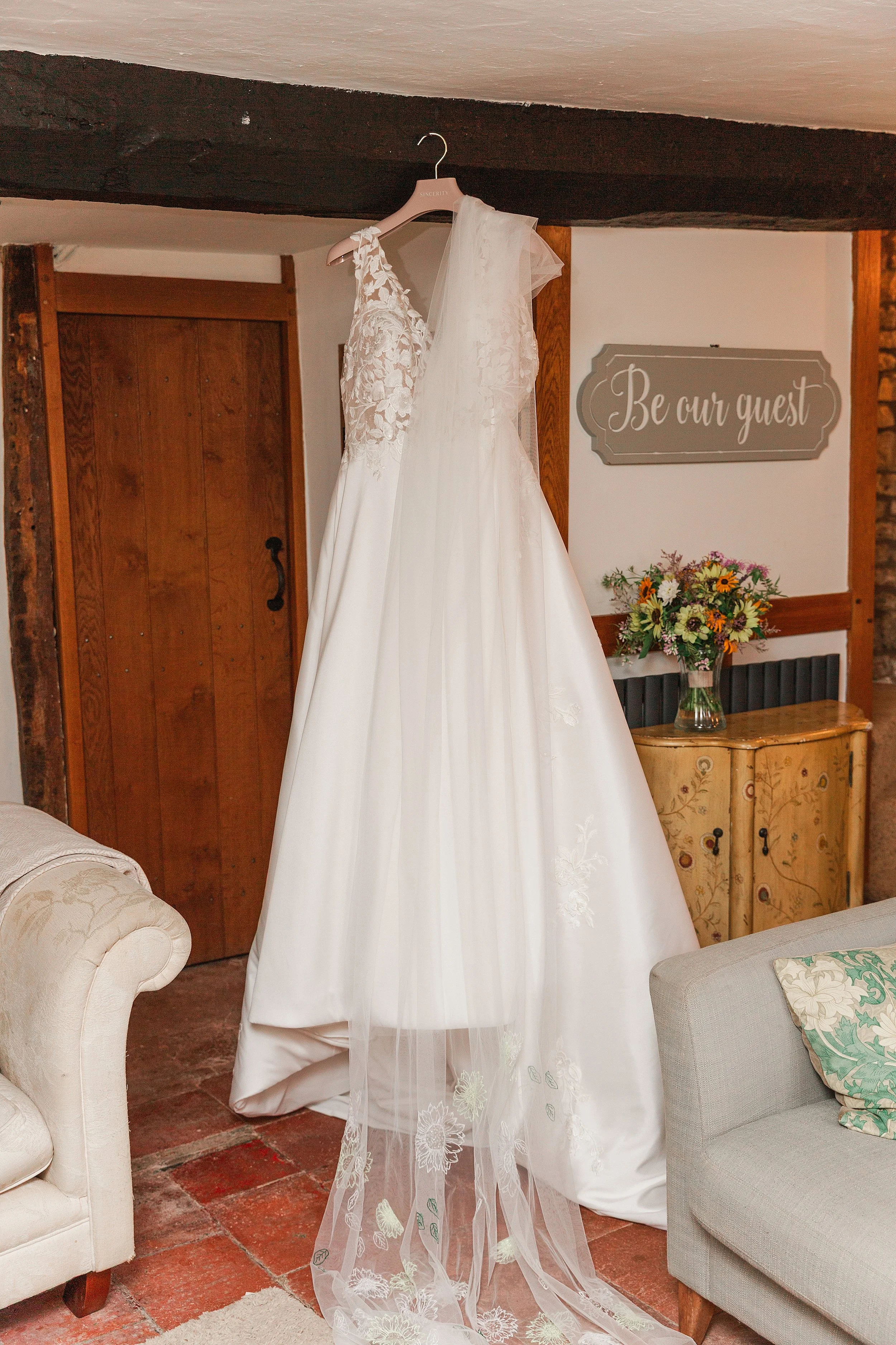 A white lace wedding dress hanging from a dark wooden beam in a farmhouse with a sign on the wall that says be my guest