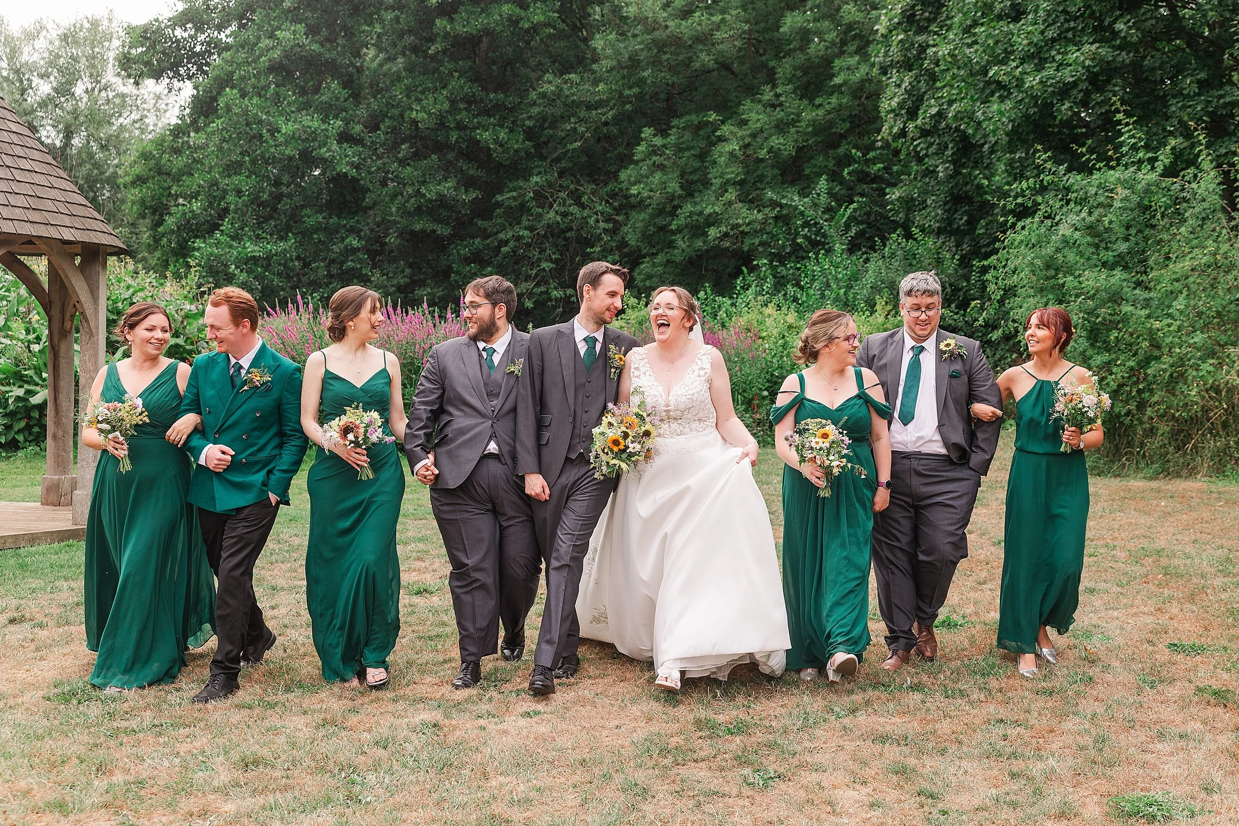 A bride with brown hair and glasses laughing with her groom with dark hair and grey suit whilst walking with their bridesmaids in green dresses and groomsman