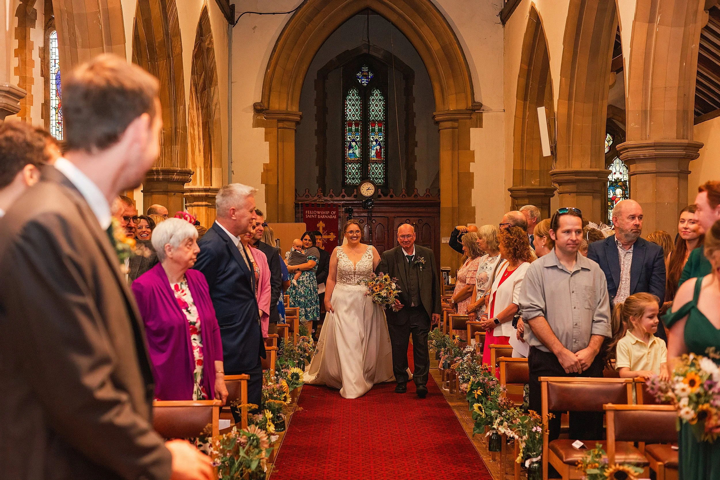 A bride walking down the aisle with her father whilst her groom watches her, there is a red carpet on the floor