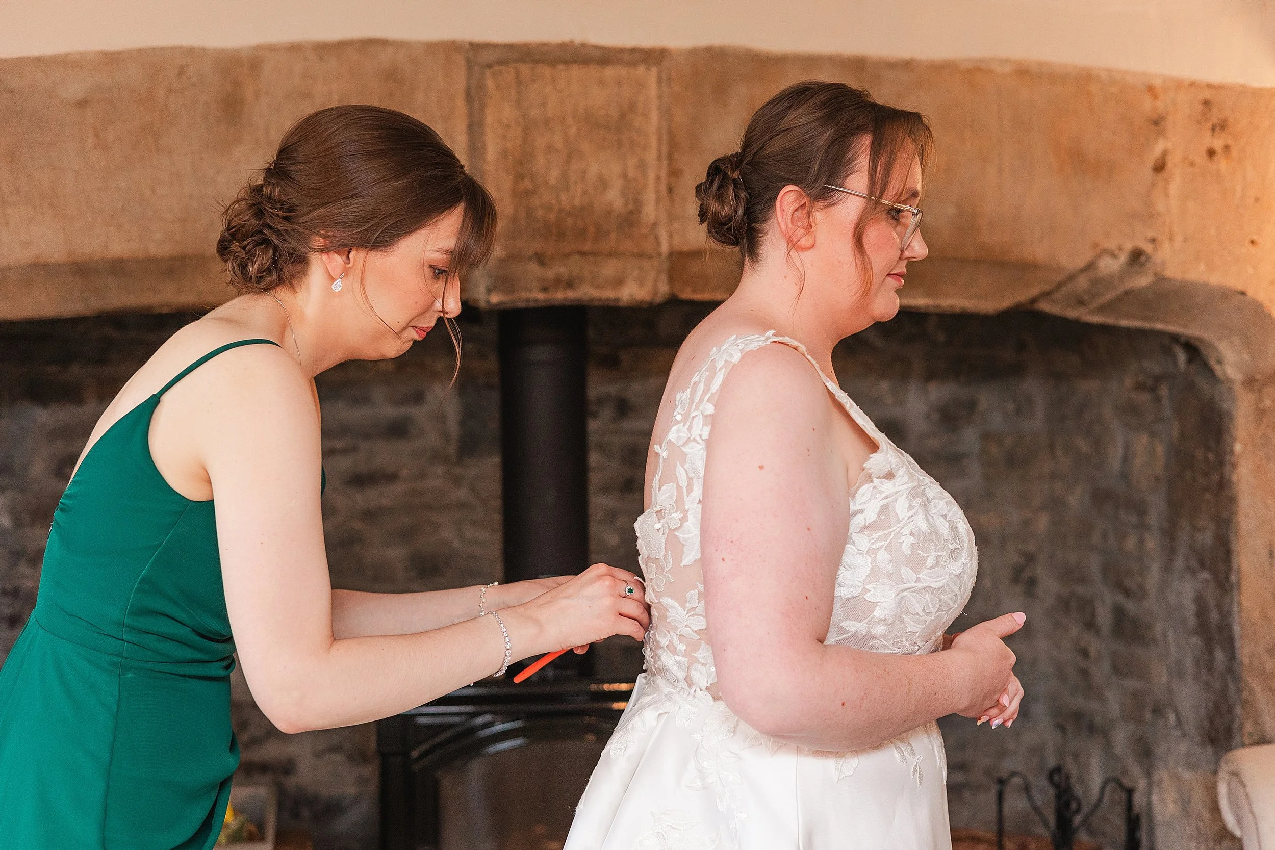 A bridesmaid with dark hair wearing an emerals green dress doing up the buttons on brides dress standing in front of a farmhouse fireplace