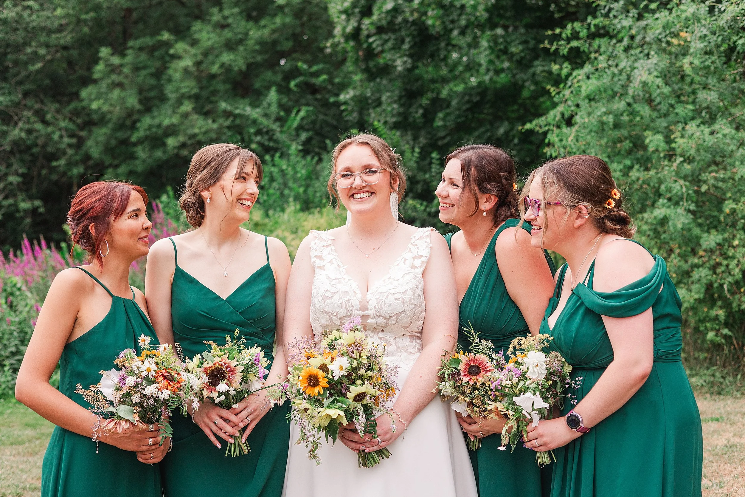 A bride with brown hair and glassed smiling at the camera standing with her 4 bridesmaids dressed in forrest green dresses smiling at her