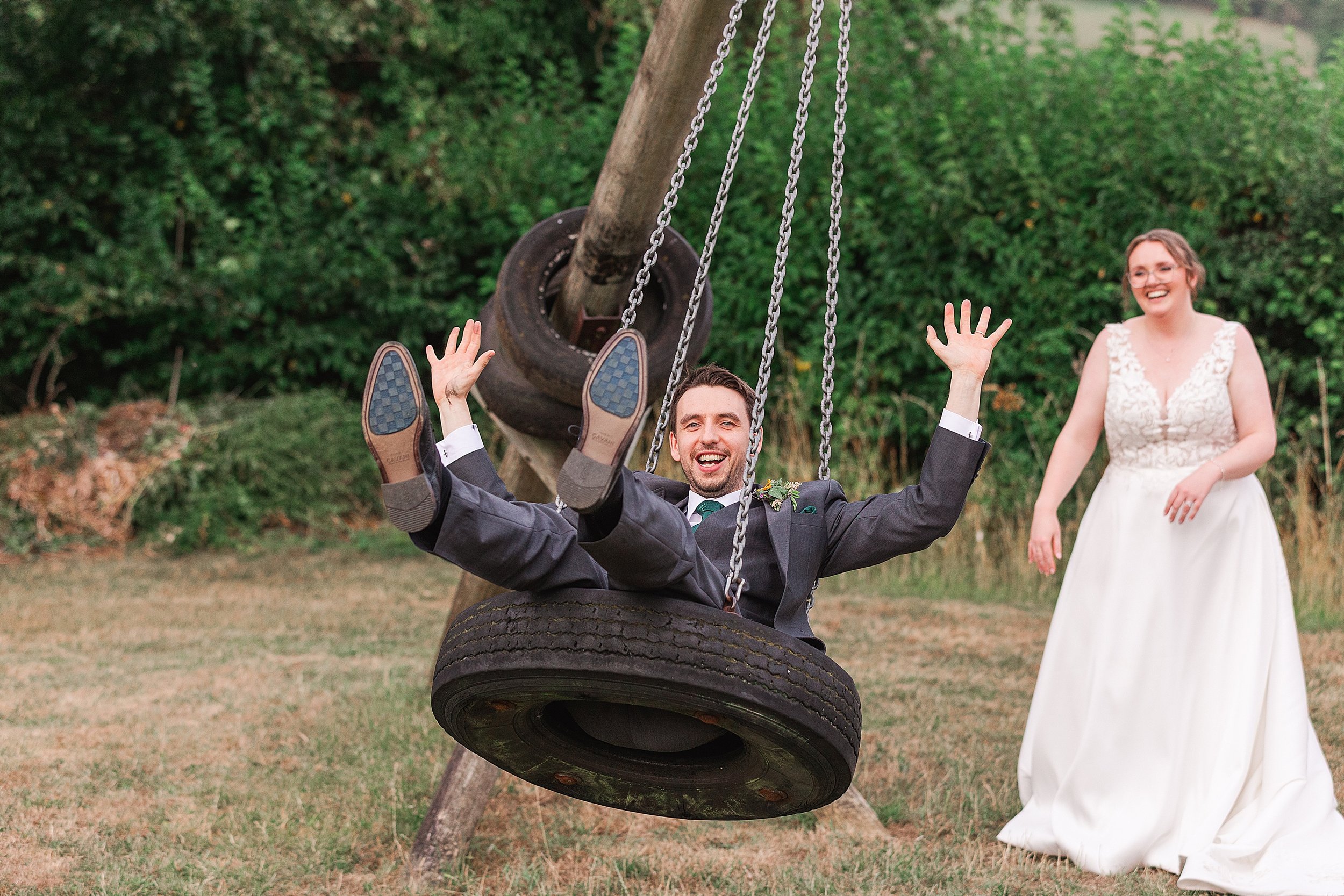 A groom dressed in grey suit swinging on a tyre whilst laughing with his hands in the air