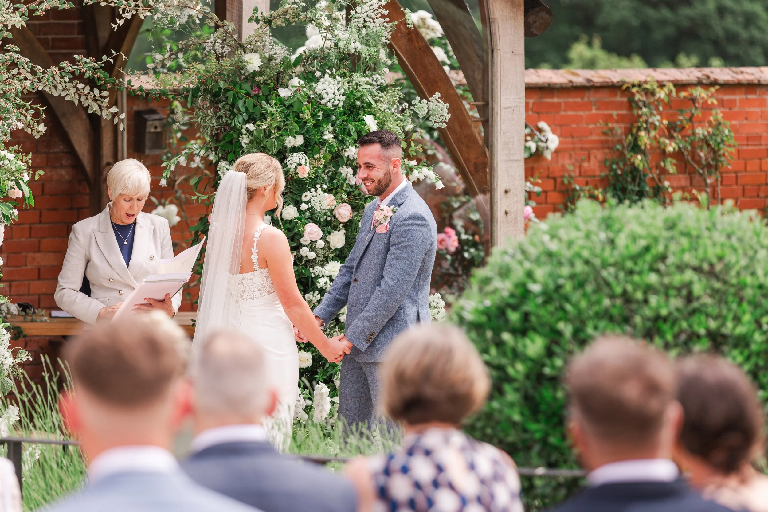 A groom dressed in a blue suit smiling at his bride during their outdoor wedding ceremony